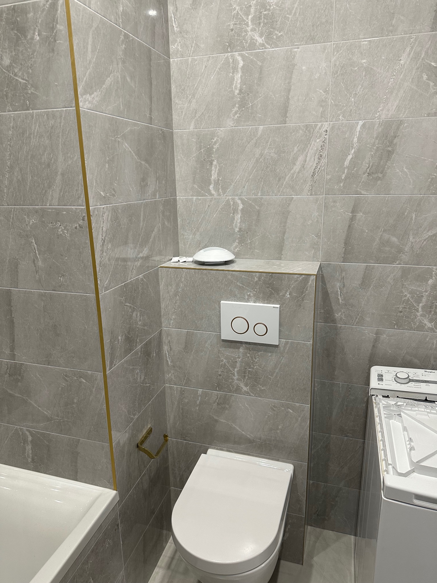 Wykończona łazienka z szarymi płytkami imitującymi marmur, złotymi akcentami, podwieszaną toaletą i pralką. Minimalistyczny design, nowoczesne wykończenie.
