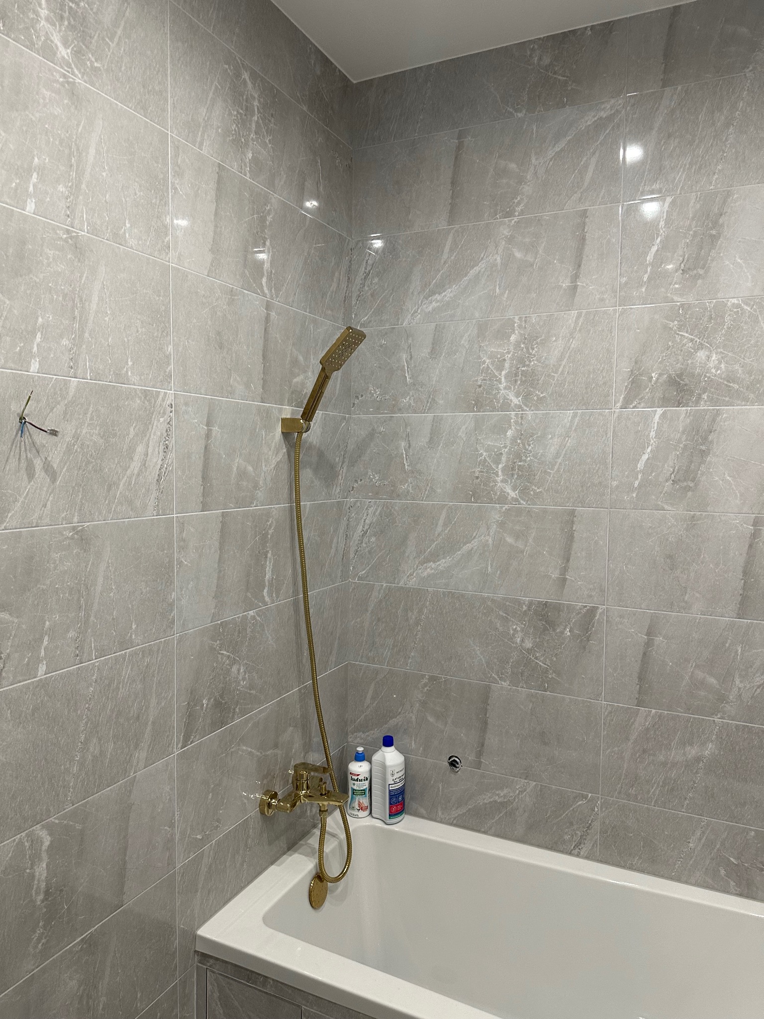 Wykończona łazienka z wanną, szarymi płytkami imitującymi marmur i złotą armaturą. Widoczne butelki z płynami do kąpieli. Minimalistyczny design.