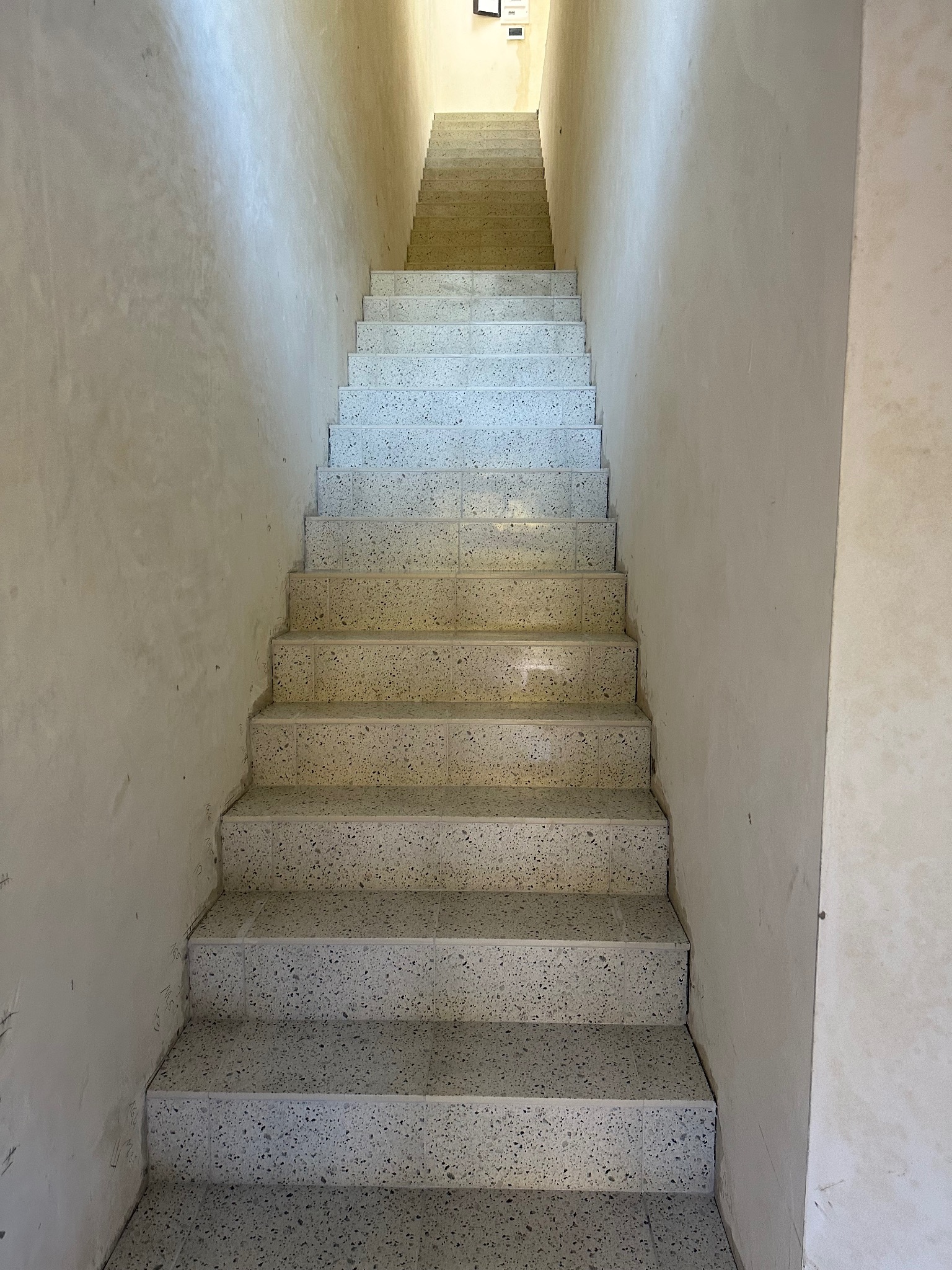 Klatka schodowa z terazzo, widok z dołu. Ściany w jasnym odcieniu beżu, schody w odcieniach szarości. Ujęcie centralne, symetryczne, z widocznym oświetleniem na górze.