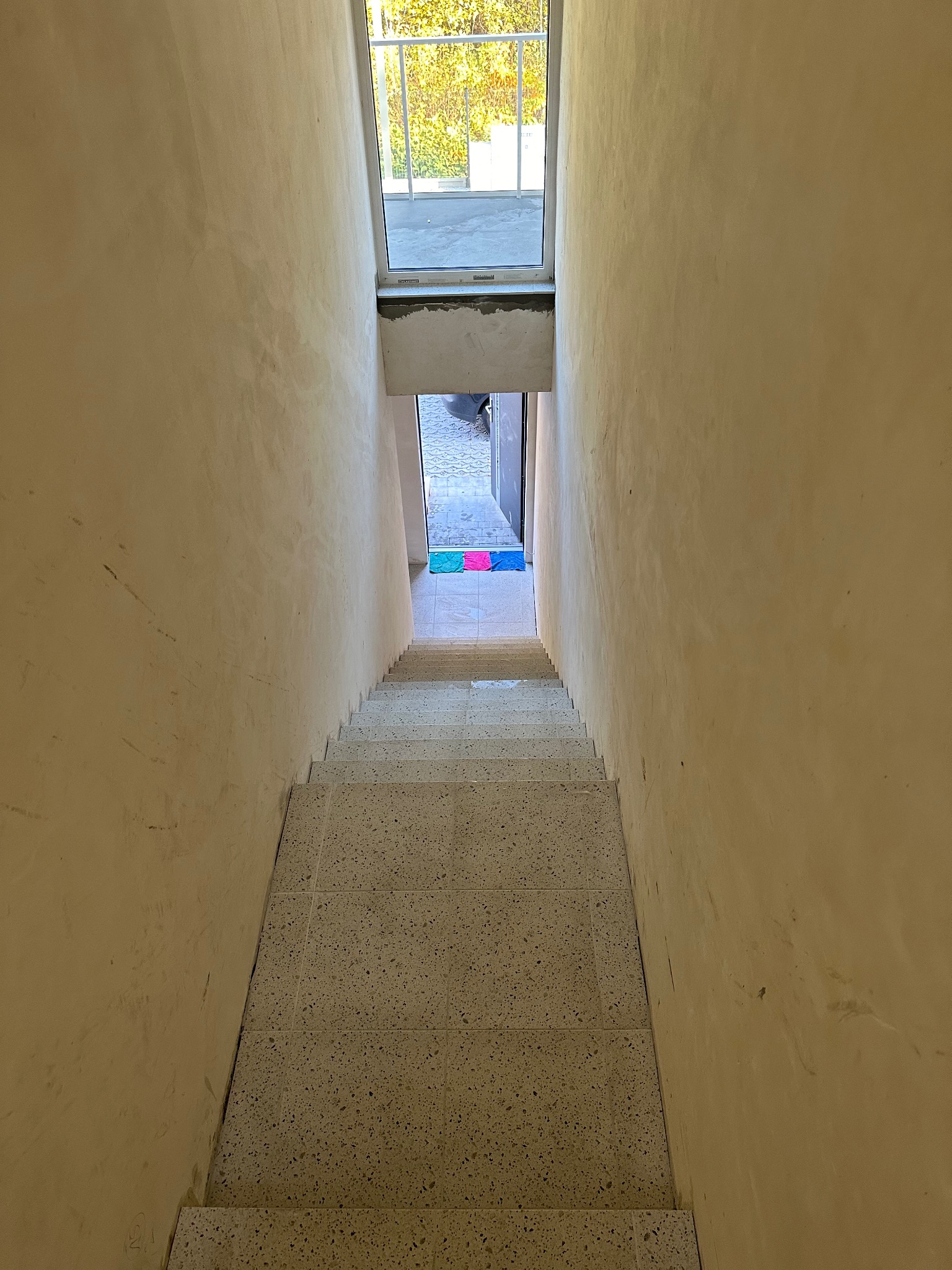 Klatka schodowa w trakcie prac wykończeniowych: ściany po szpachlowaniu, schody wyłożone płytkami z terazzo, widok z góry na dół, w tle okno i wejście.