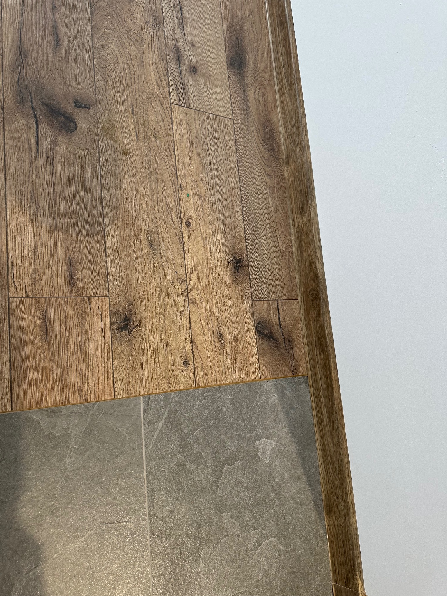 Połączenie podłogi: panele drewnopodobne i szare płytki. Widoczna biała ściana i drewniana listwa przypodłogowa. Ujęcie z góry, ukazujące detale materiałów.