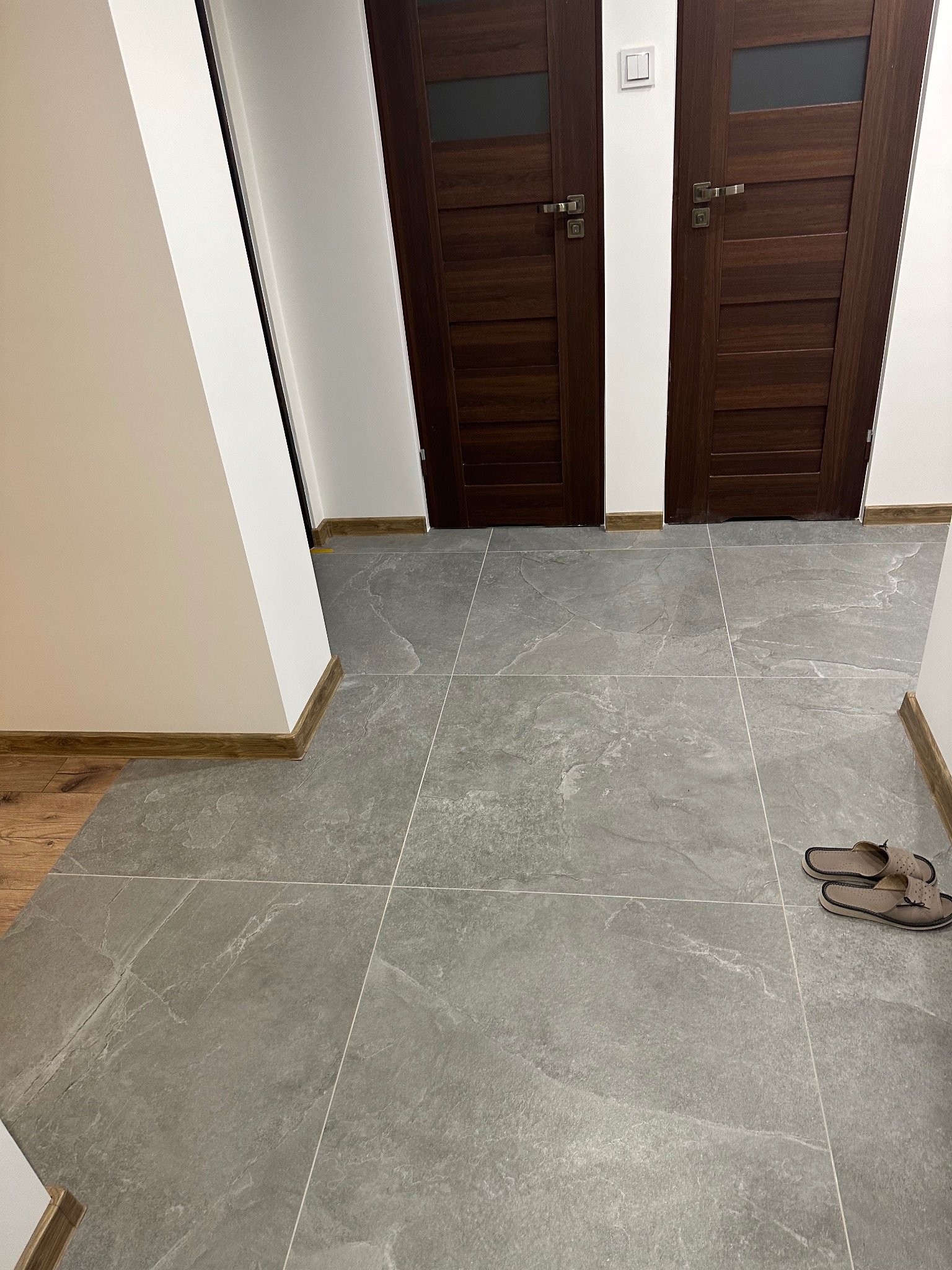 Szare, kwadratowe płytki podłogowe imitujące kamień w korytarzu, z drewnianymi listwami przypodłogowymi i dwiema parami drzwi z ciemnego drewna.