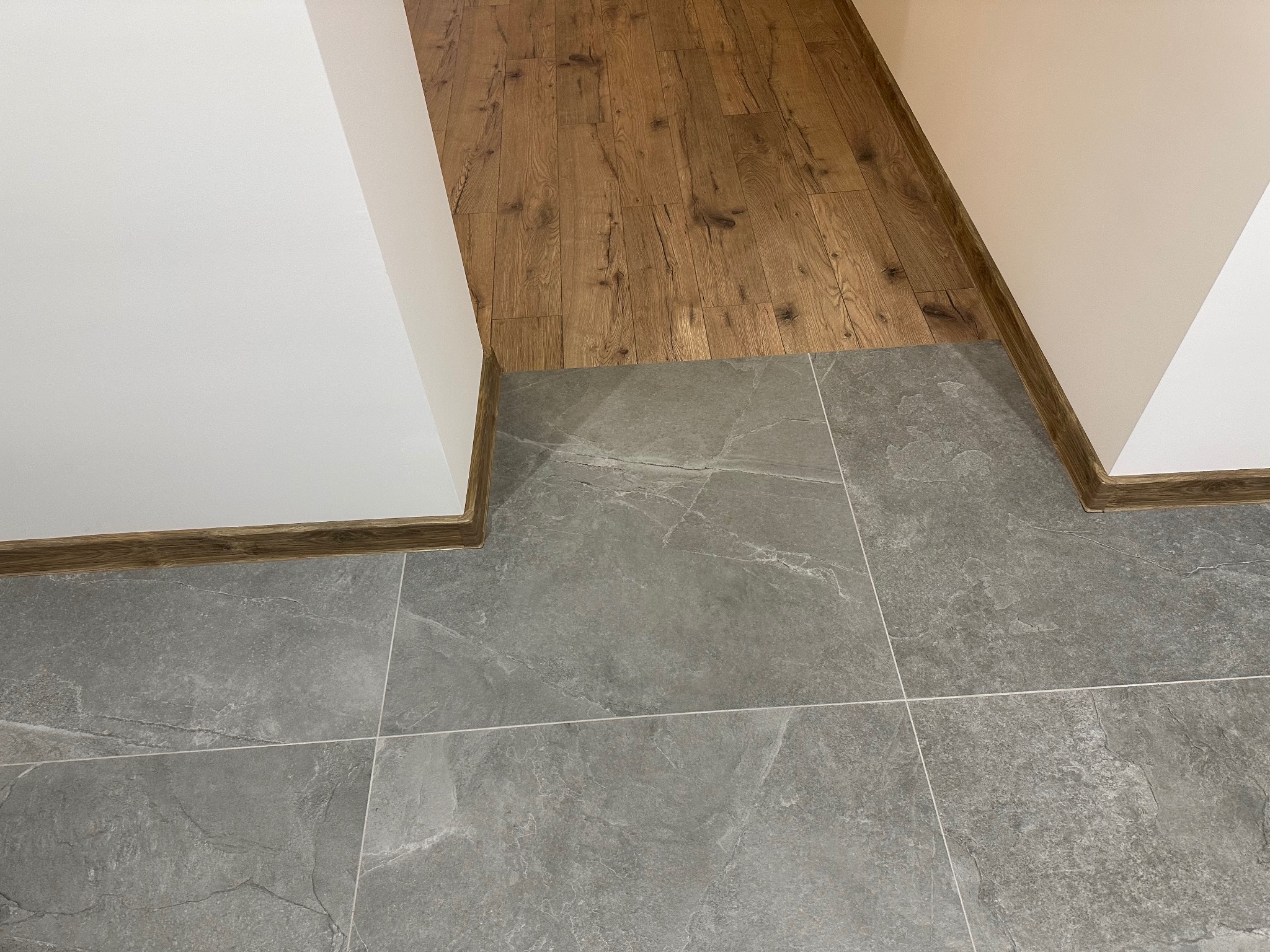Połączenie podłogi drewnianej z szarą, kamienną posadzką w korytarzu. Widoczne listwy przypodłogowe w kolorze drewna. Ujęcie z góry, podkreślające fakturę materiałów.