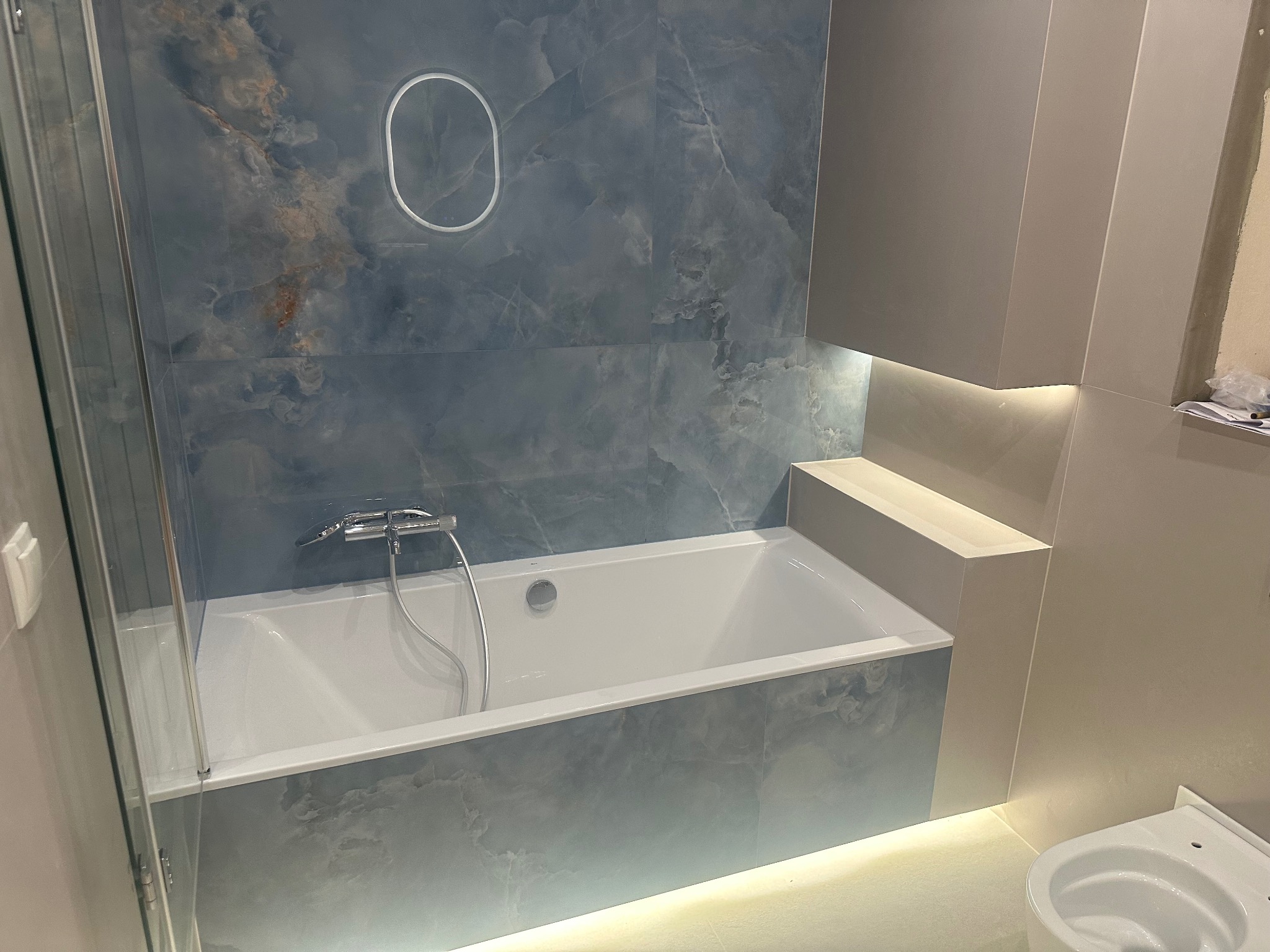 Nowoczesna łazienka z wanną obudowaną płytkami imitującymi marmur, podświetleniem LED i szklanym parawanem nawannowym. Minimalistyczny design, eleganckie wykończenie.