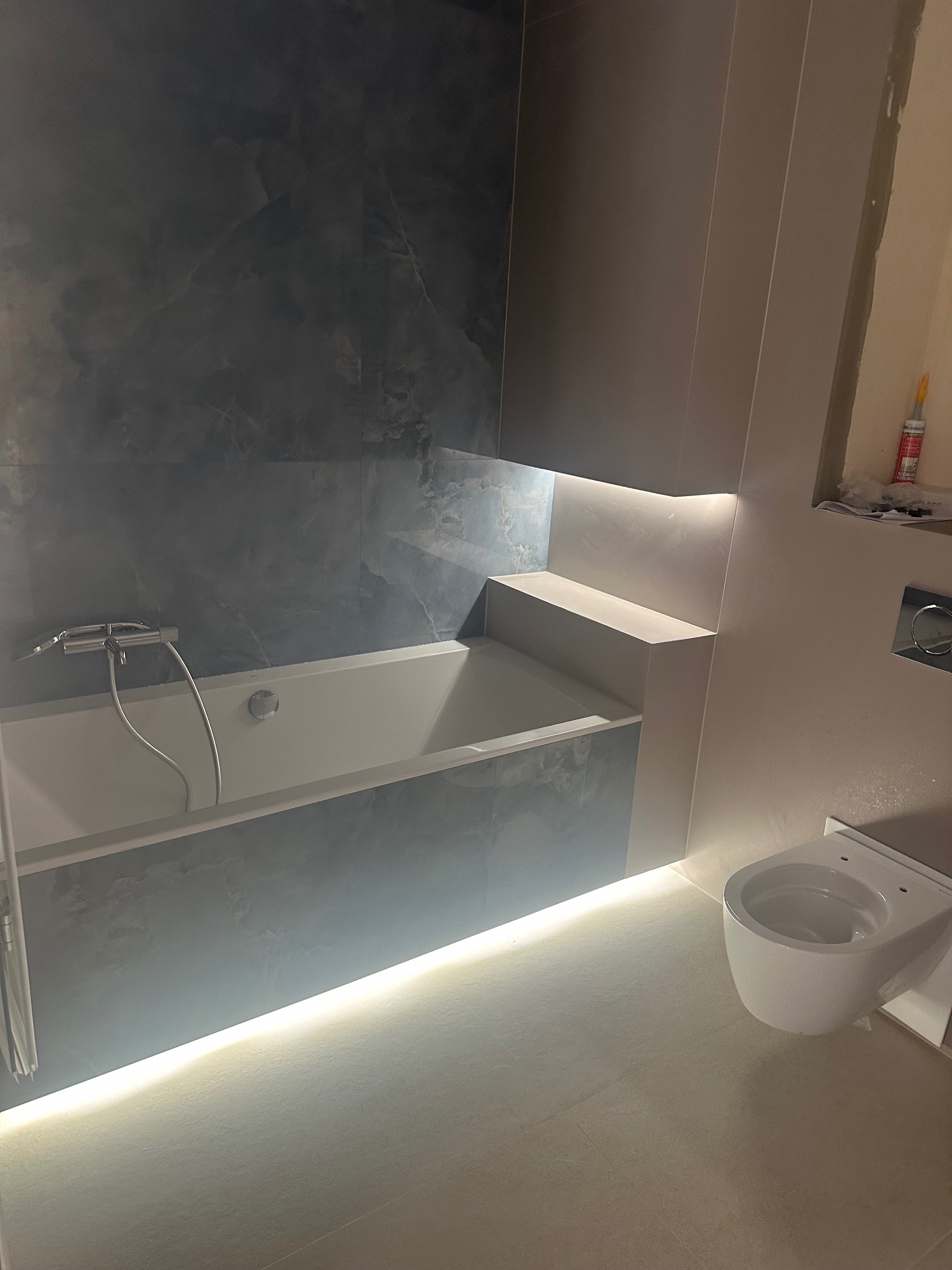 Nowoczesna łazienka z podświetlaną wanną w zabudowie i wiszącą toaletą. Ściany wykończone płytkami imitującymi marmur. Oświetlenie LED podkreśla minimalistyczny design.