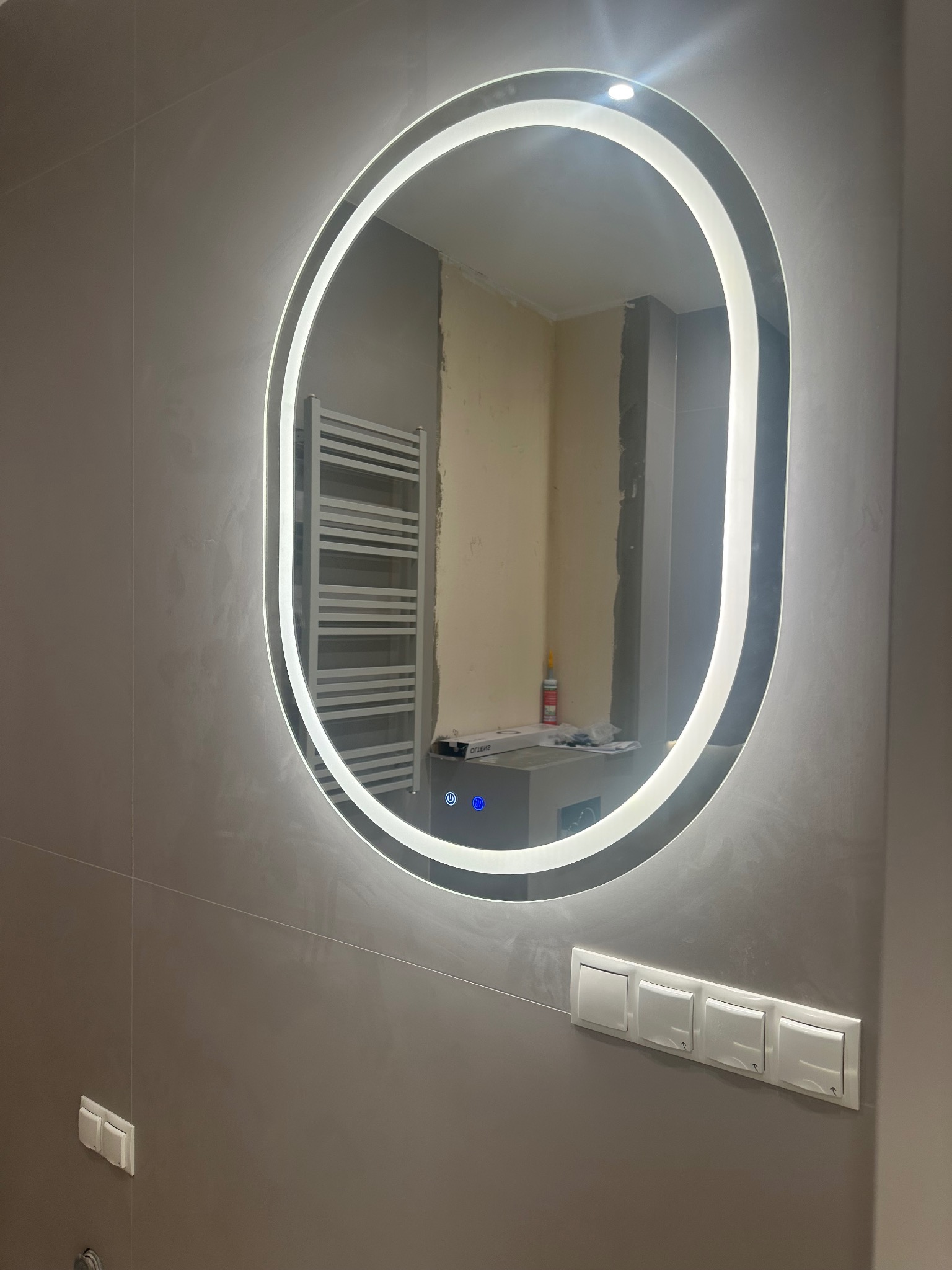 Owalne lustro z podświetleniem LED w wykończonej łazience, odbijające fragment remontowanego wnętrza z grzejnikiem drabinkowym i akcesoriami. Szare płytki i białe włączniki.