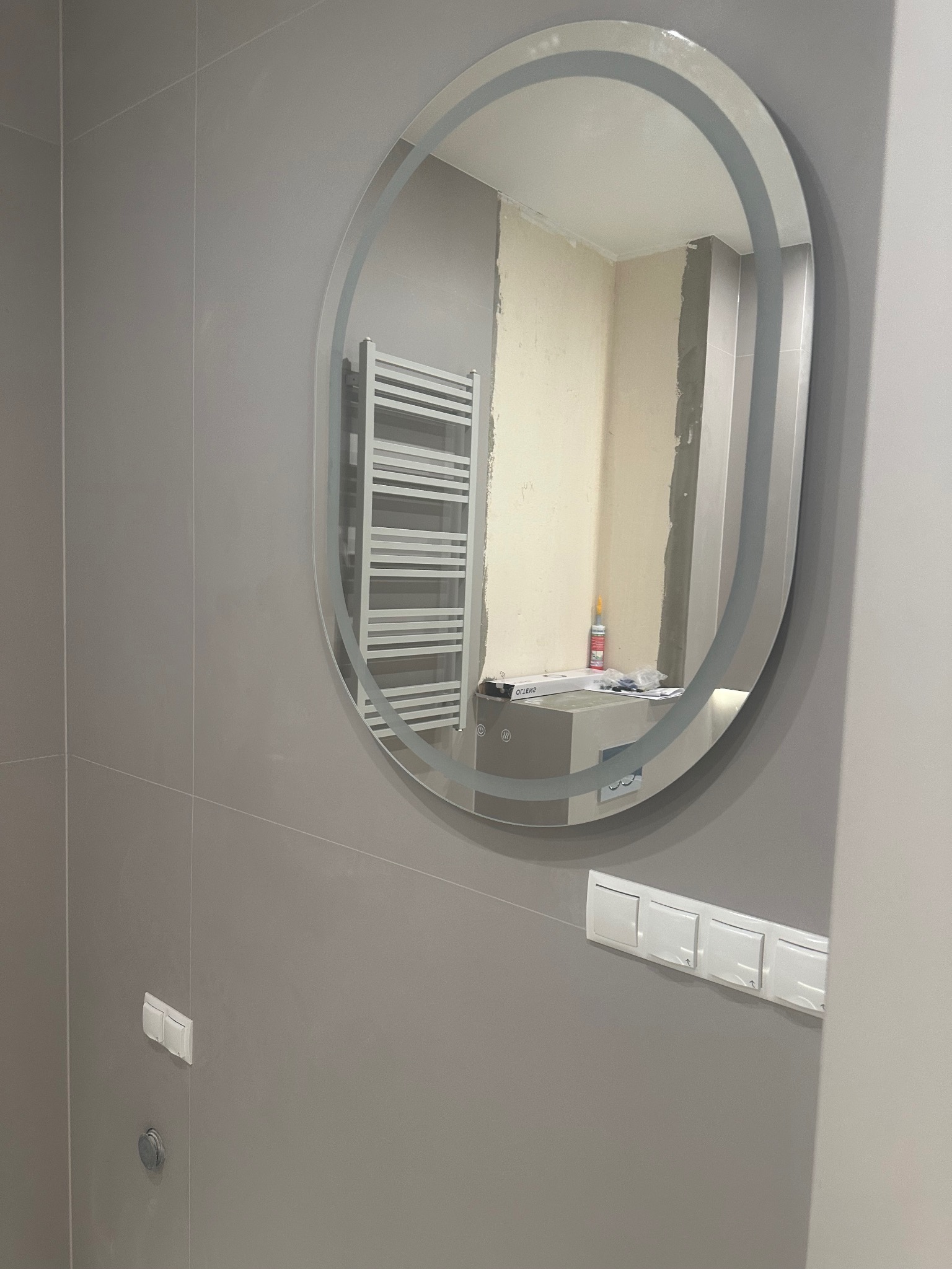 Szare płytki w łazience z okrągłym lustrem LED, w którym odbija się grzejnik i fragment ściany w trakcie prac wykończeniowych. Widoczne włączniki światła.
