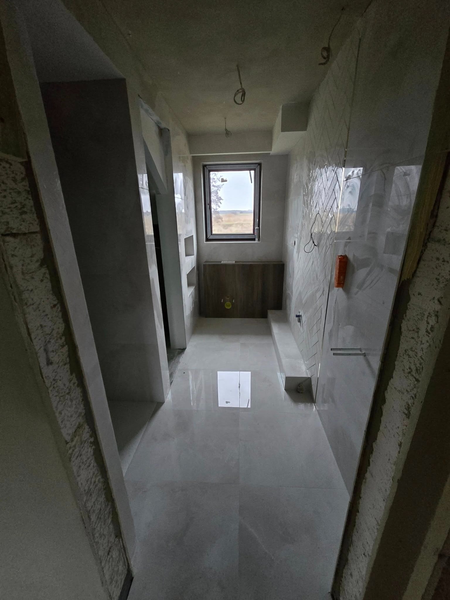 Wnętrze łazienki w trakcie wykańczania: położone płytki podłogowe i ścienne, widoczne instalacje, okno z widokiem na krajobraz. Surowy strop i ściany w trakcie prac.