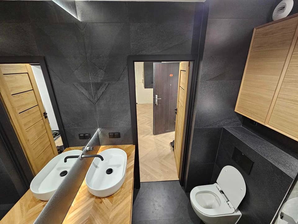 Nowoczesna łazienka z czarnymi płytkami, drewnianymi szafkami i blatem pod umywalki ułożonym w jodełkę. Widoczne dwie białe umywalki i toaleta.