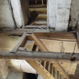 Mann sp. z o.o. - Surowe drewniane elementy konstrukcyjne w starym budynku, widok z dołu. Fragmenty ścian z widocznymi śladami czasu i instalacji. W tle widoczne schody i otwór w podłodze.