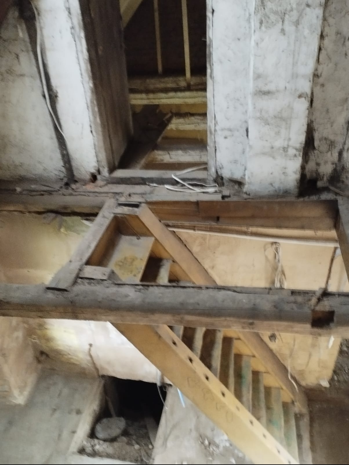 Surowe drewniane elementy konstrukcyjne w starym budynku, widok z dołu. Fragmenty ścian z widocznymi śladami czasu i instalacji. W tle widoczne schody i otwór w podłodze.