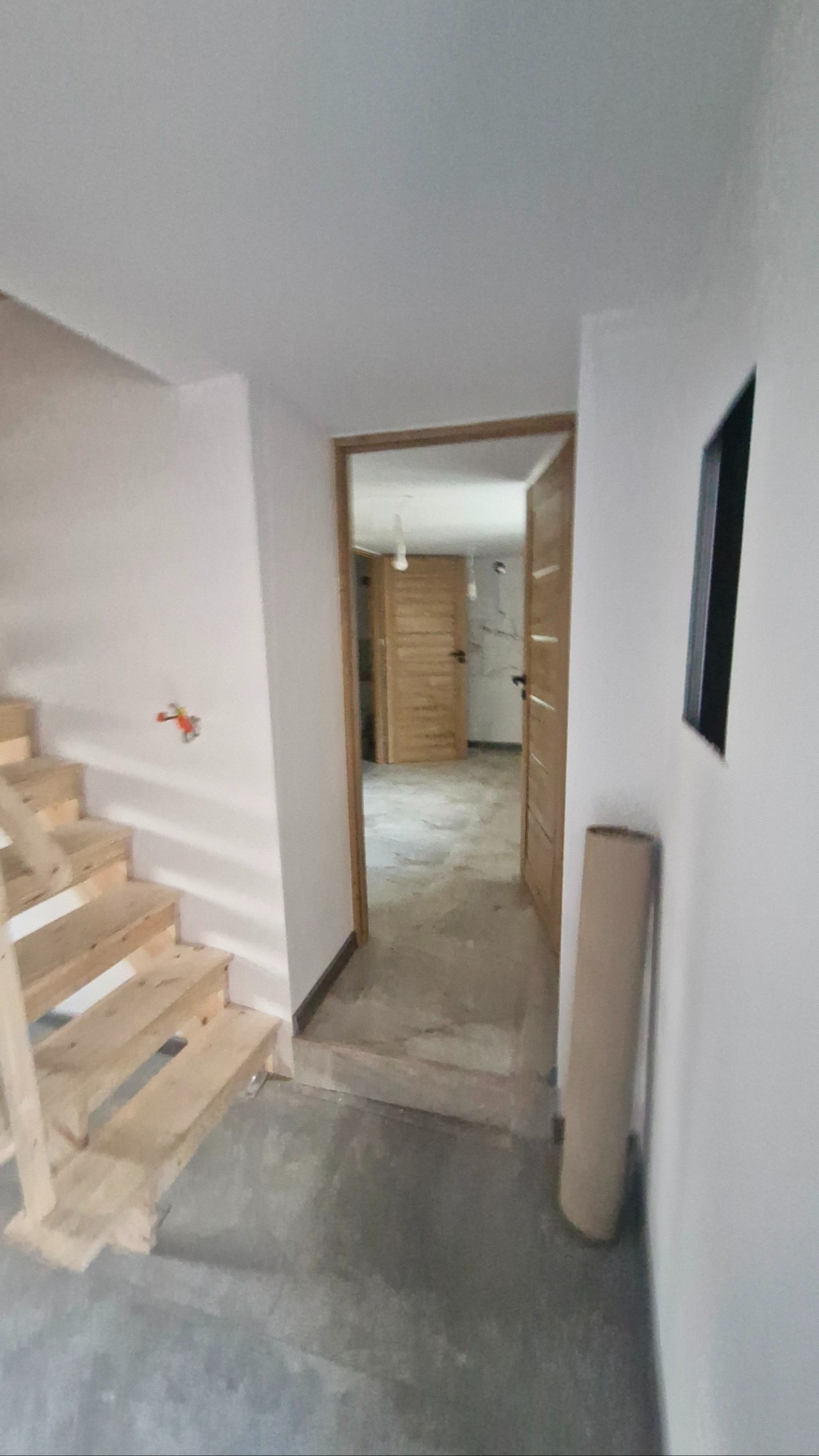 Wnętrze w trakcie wykończenia: surowe schody drewniane, białe ściany z widoczną instalacją elektryczną, otwarte drzwi ukazujące dalszą część pomieszczenia z drewnianymi drzwiami.