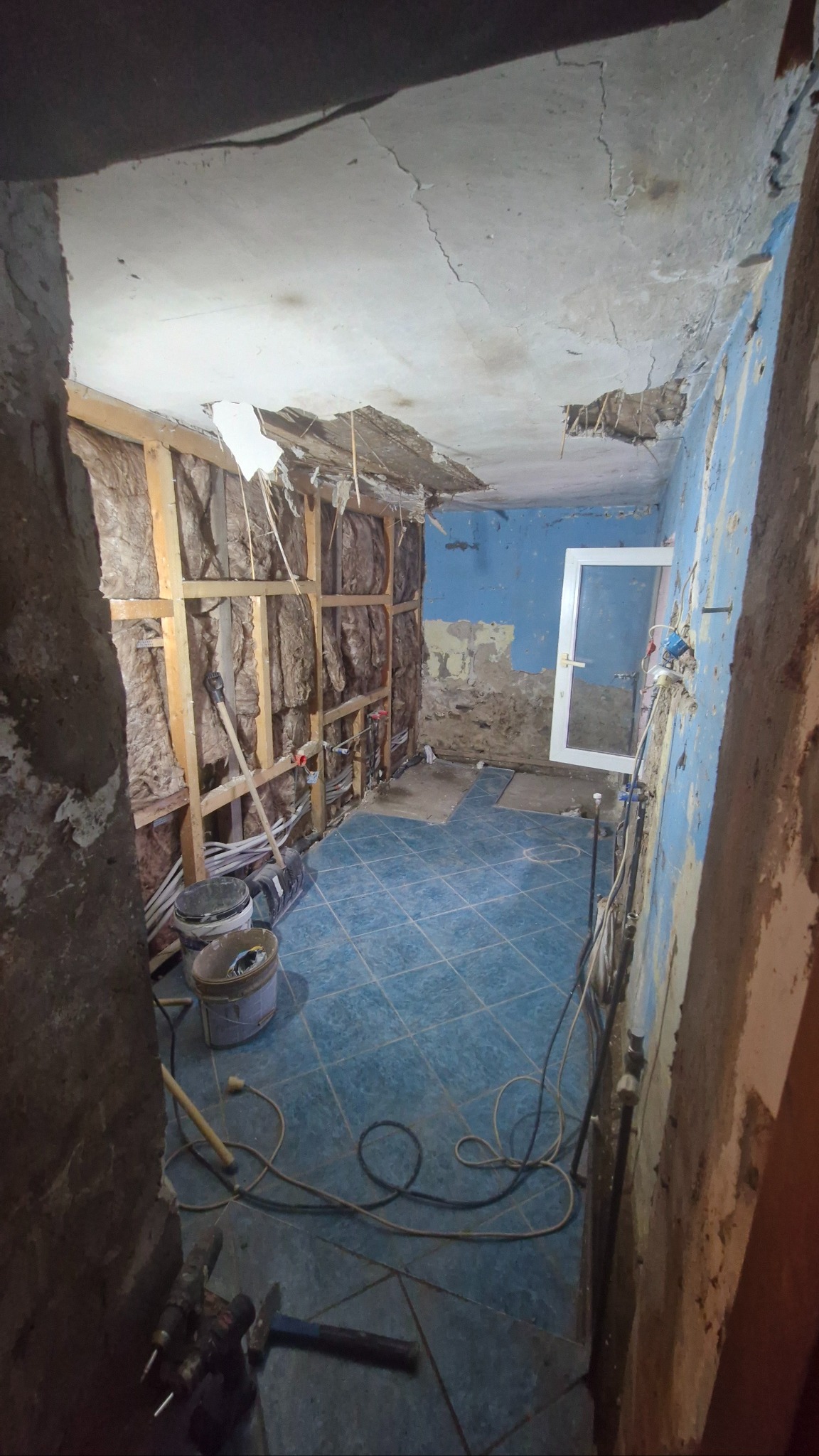 Wnętrze w trakcie remontu: odsłonięte ściany z izolacją, instalacje, niebieska podłoga z płytek, widoczne narzędzia i materiały budowlane. Prace w toku.