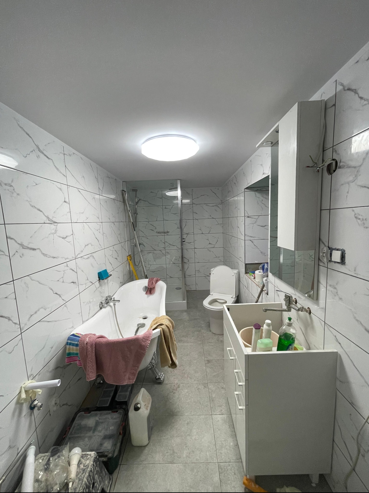 Remont łazienki w trakcie: białe marmurowe płytki, wolnostojąca wanna na srebrnych nóżkach, biała szafka z umywalką, toaleta. Widoczne narzędzia i materiały budowlane.