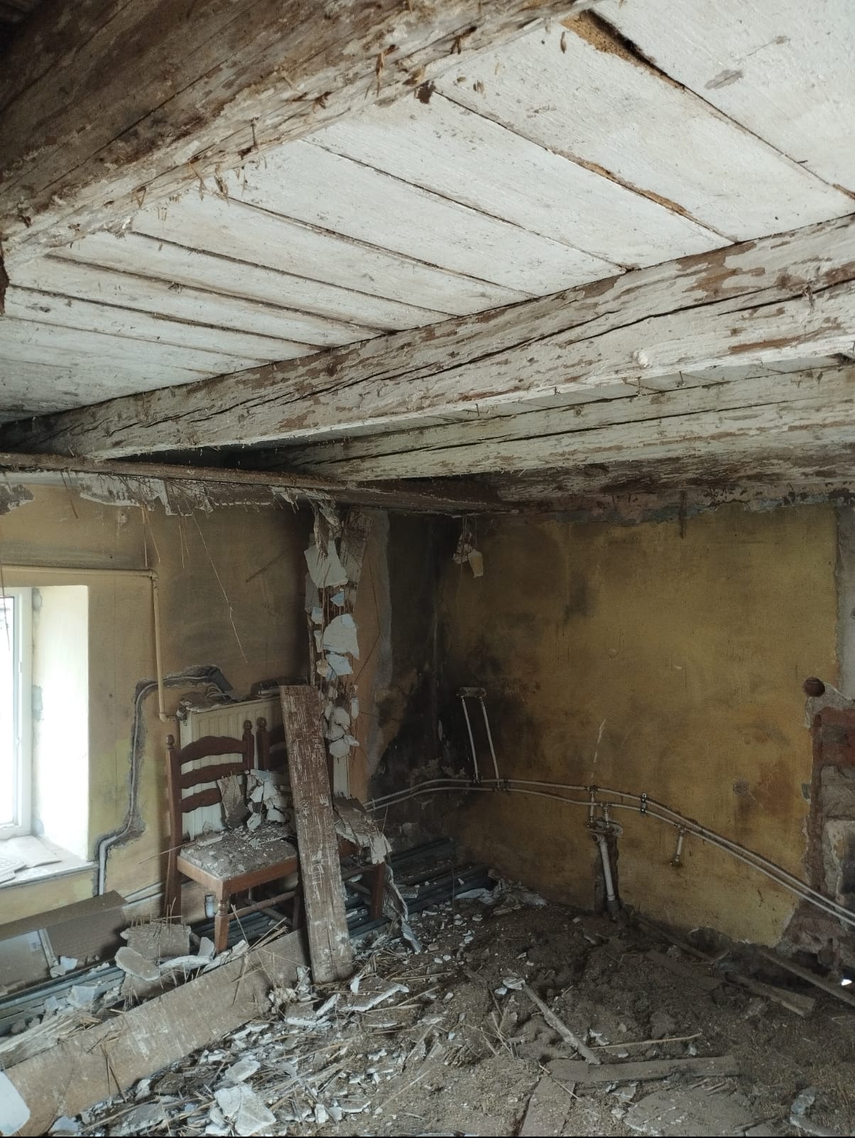Wnętrze pokoju w trakcie demontażu: odsłonięty drewniany strop, fragmenty ścian i gruzu na podłodze, stare rury. Widoczne krzesło i okno.
