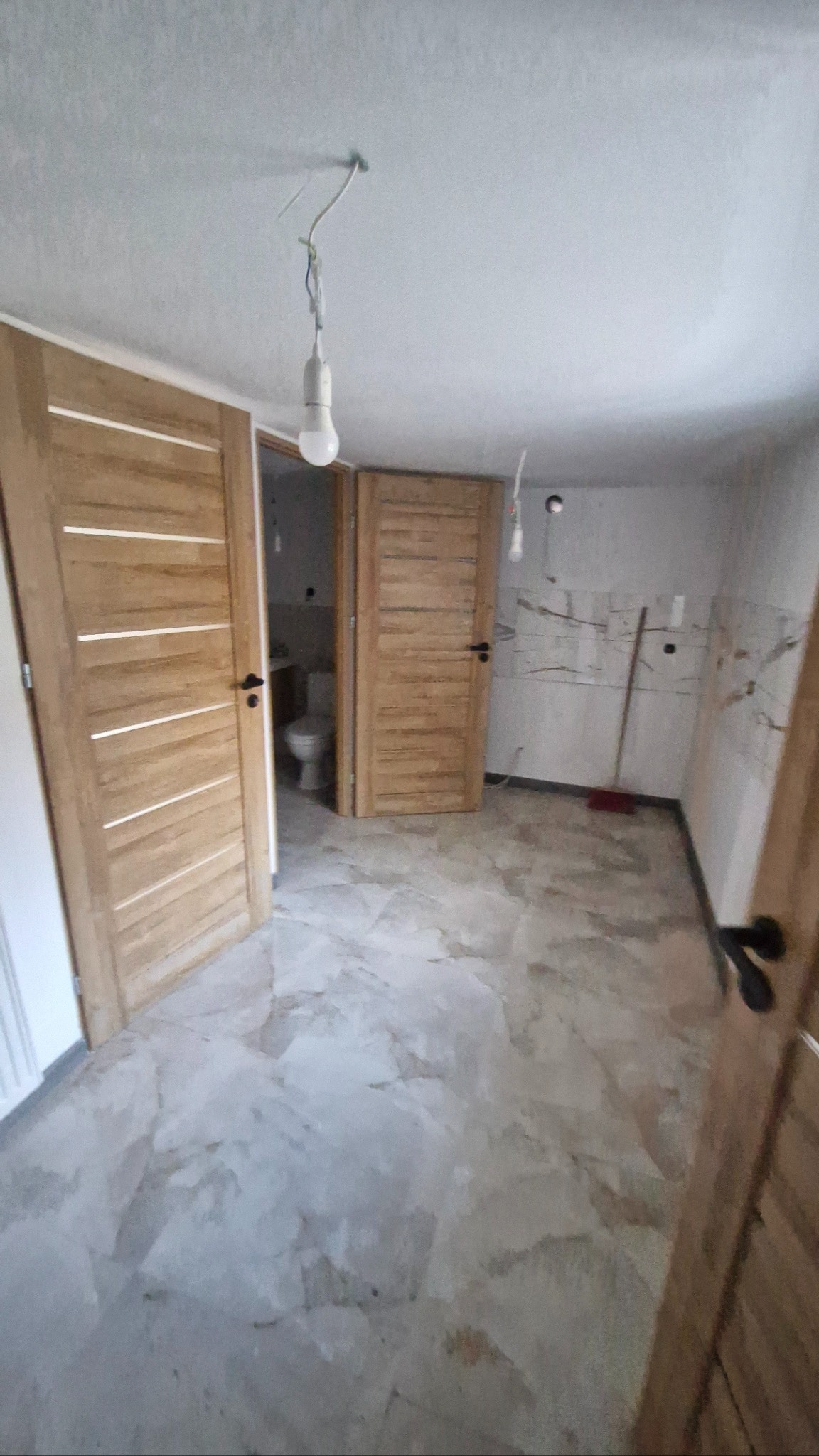 Wnętrze po remoncie: marmurowa podłoga, drewniane drzwi z poziomymi panelami, widok na toaletę. Niedokończone wykończenie ścian, wisząca żarówka.