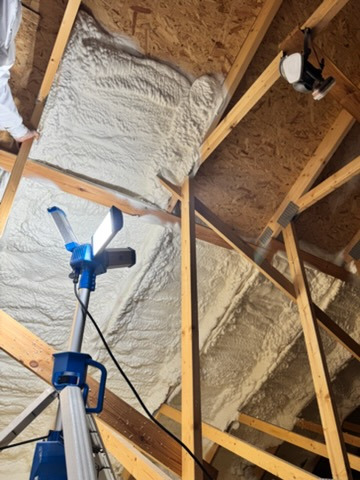 Poddasze ocieplone pianką PUR, widoczne drewniane elementy konstrukcyjne i lampa budowlana. Aplikacja pianki w trudno dostępnych miejscach.