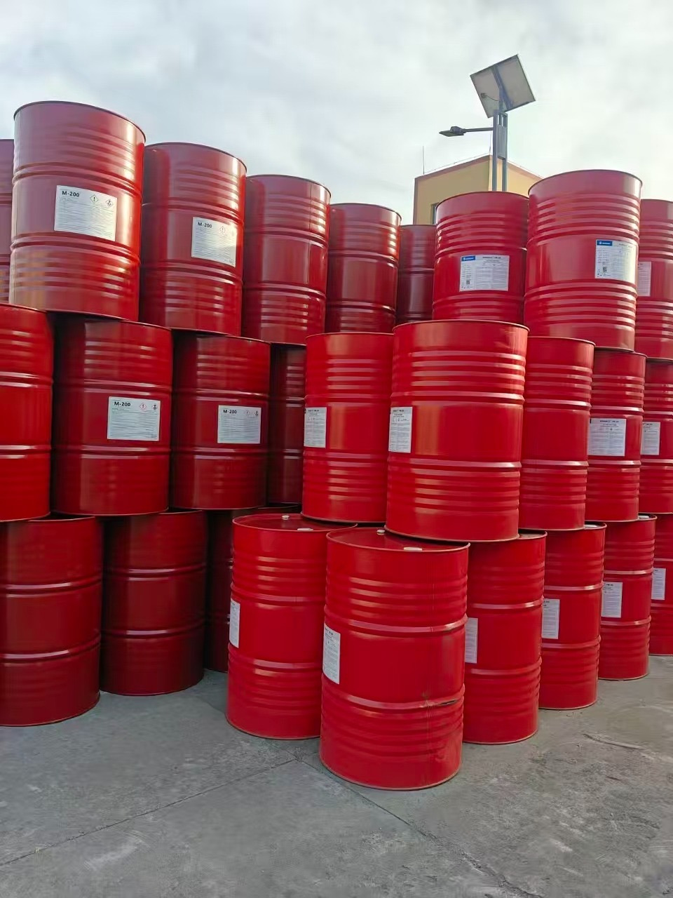 Czerwone beczki M-200 ułożone w stos, prawdopodobnie z komponentami do pianki PUR, na zewnątrz, w tle słup oświetleniowy i budynek.