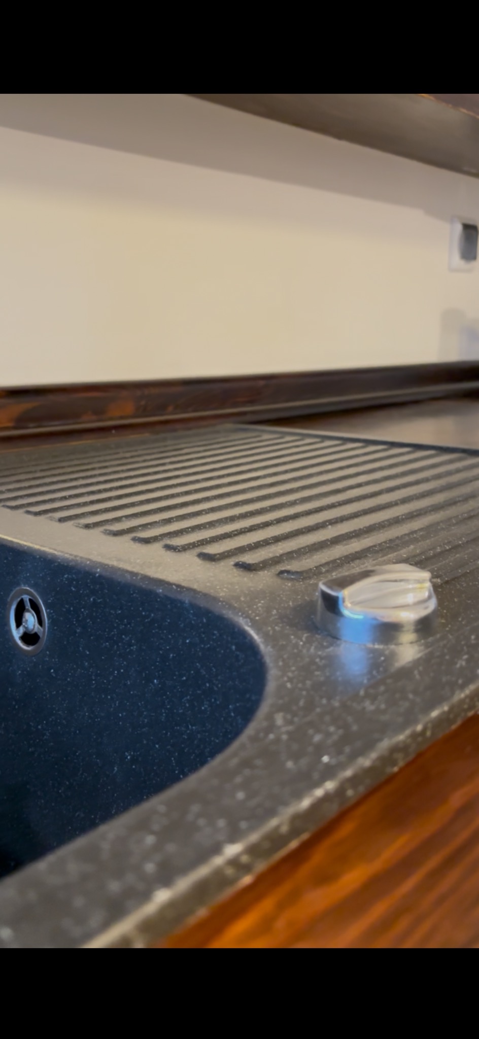 Granitowy zlew kuchenny z ociekaczem, widoczny z bliska pod kątem. Elementy armatury i drewniany blat. Powierzchnia zlew lekko zakurzona.