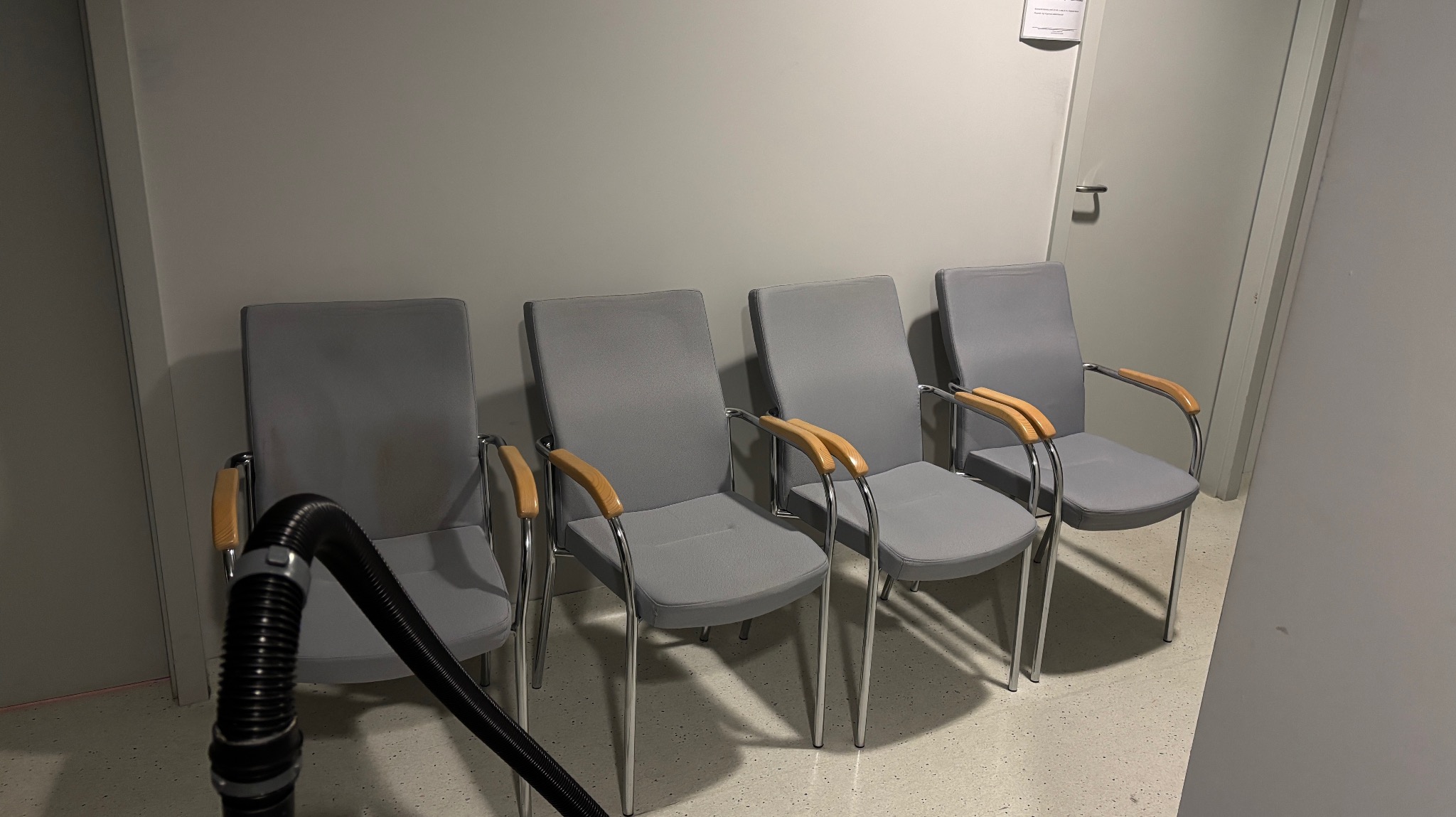 Szereg czterech szarych krzeseł z drewnianymi podłokietnikami, czyszczonych odkurzaczem piorącym. Minimalistyczne wnętrze, jasne ściany i podłoga. Widoczny fragment rury odkurzacza.