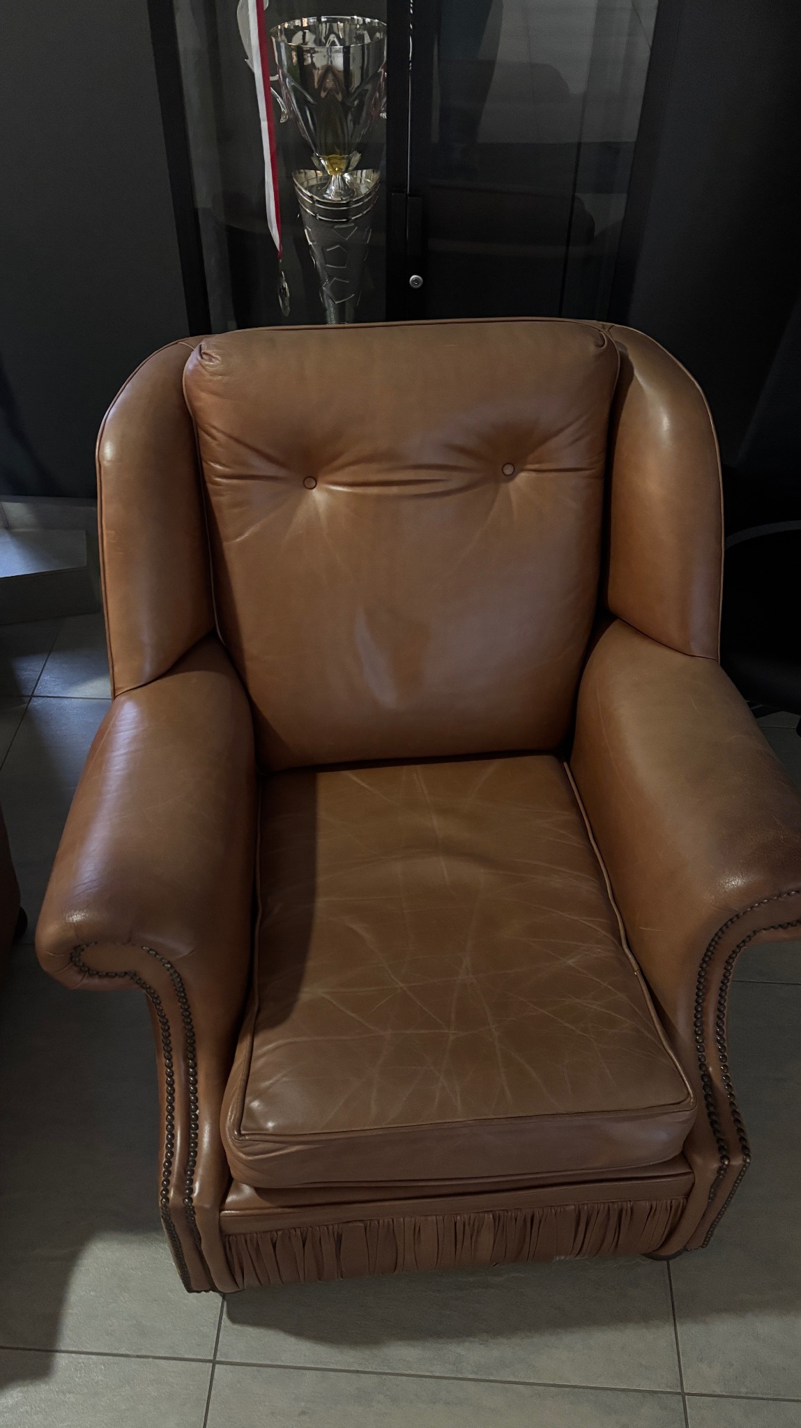 Brązowy, skórzany fotel typu uszak z dekoracyjnymi nitami i marszczeniem u dołu, stojący na szarej podłodze, z trofeum widocznym w tle.