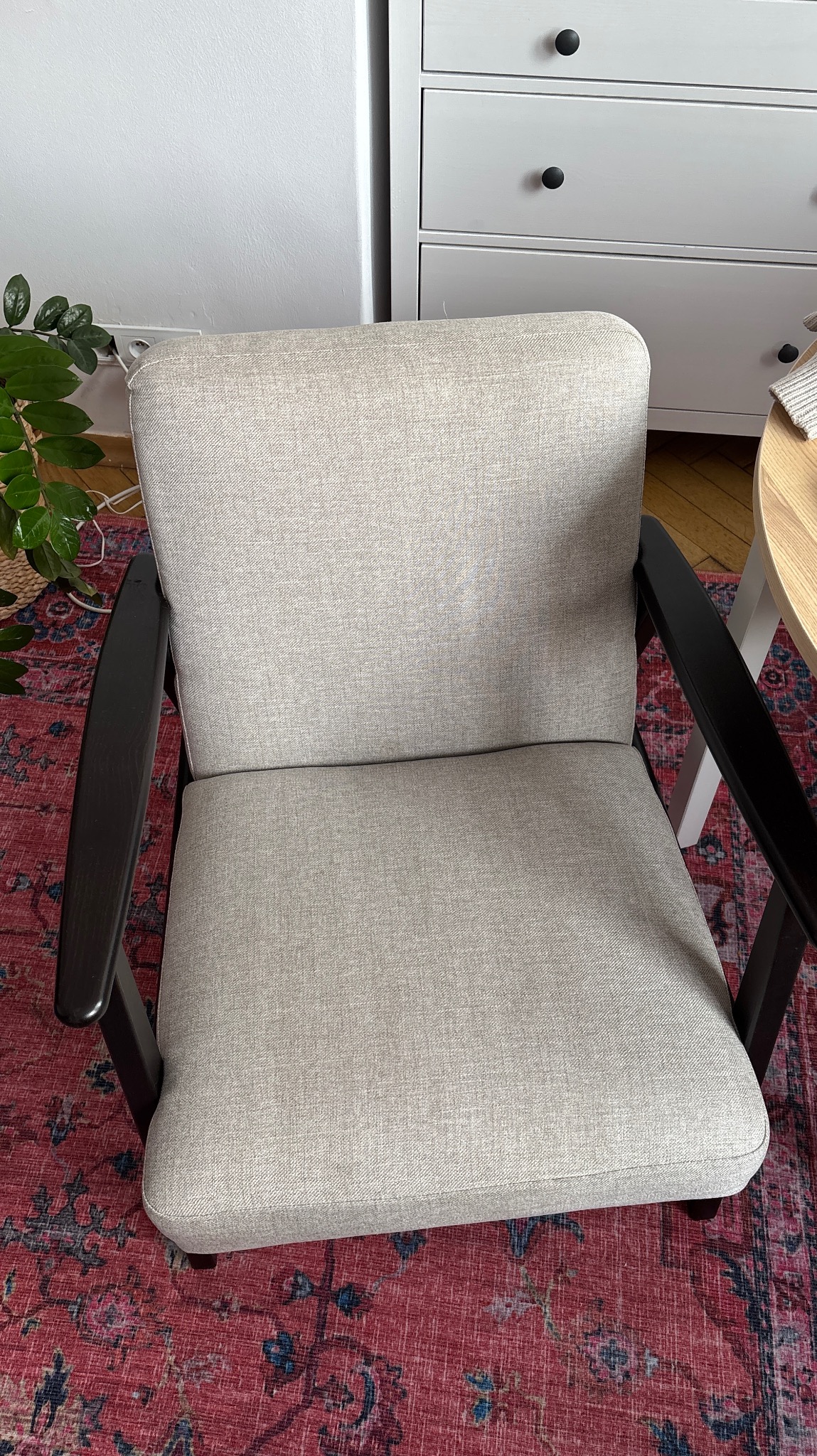 Beżowy fotel tapicerowany z ciemnymi podłokietnikami na wzorzystym, czerwonym dywanie. W tle biała komoda i fragment rośliny doniczkowej.