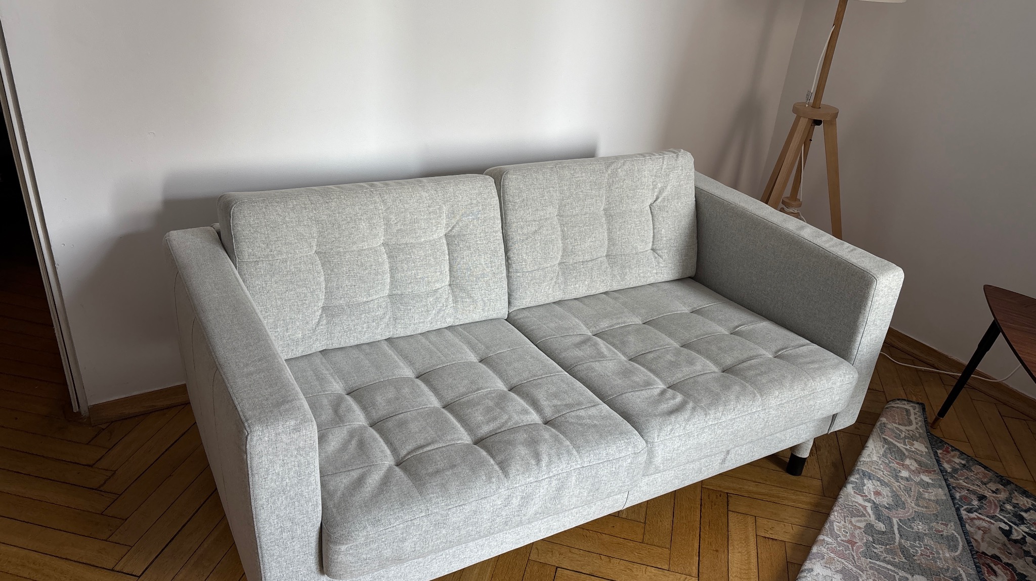 Jasnoszara, dwuosobowa sofa z pikowanymi siedziskami na drewnianej podłodze, obok lampa i stolik. Minimalistyczny styl, neutralne kolory.