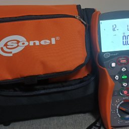Pomarańczowa torba narzędziowa Sonel obok miernika elektrycznego MPI-507. Widoczny wyświetlacz miernika z wartościami pomiarowymi. Sprzęt do pomiarów elektrycznych.