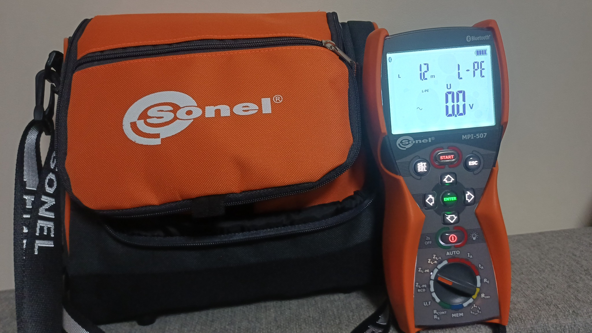 Pomarańczowa torba narzędziowa Sonel obok miernika elektrycznego MPI-507. Widoczny wyświetlacz miernika z wartościami pomiarowymi. Sprzęt do pomiarów elektrycznych.