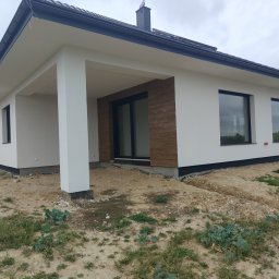 Krzysztof Samborski Docieplenia i elewacje - Nowoczesny dom jednorodzinny z jasną elewacją, ciemnym dachem i dużymi oknami. Widoczny fragment tarasu z kolumną. Teren wokół domu w trakcie zagospodarowania.