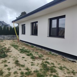 Krzysztof Samborski Docieplenia i elewacje - Biała elewacja domu jednorodzinnego z czarnymi oknami w Mełgwi. Widoczny fragment ogrodu i sąsiedni budynek. Ujęcie z boku, w pochmurny dzień.