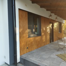 Krzysztof Samborski Docieplenia i elewacje - Nowoczesna elewacja domu z drewnianą okładziną i szarą stolarką okienną. Widoczny fragment tarasu z meblami ogrodowymi i jesienny krajobraz w tle.