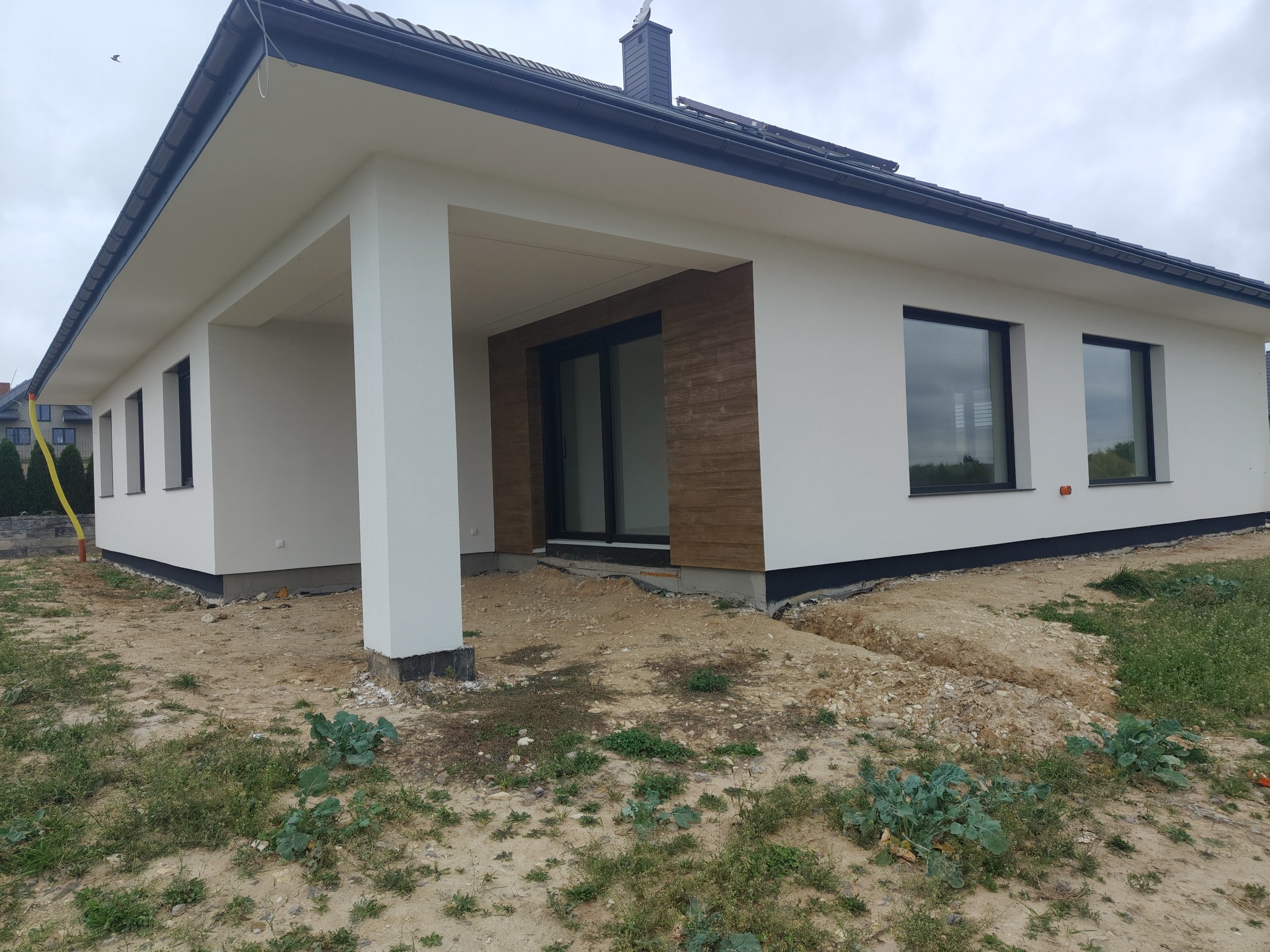 Nowoczesny dom jednorodzinny z jasną elewacją, ciemnym dachem i dużymi oknami. Widoczny fragment tarasu z kolumną. Teren wokół domu w trakcie zagospodarowania.
