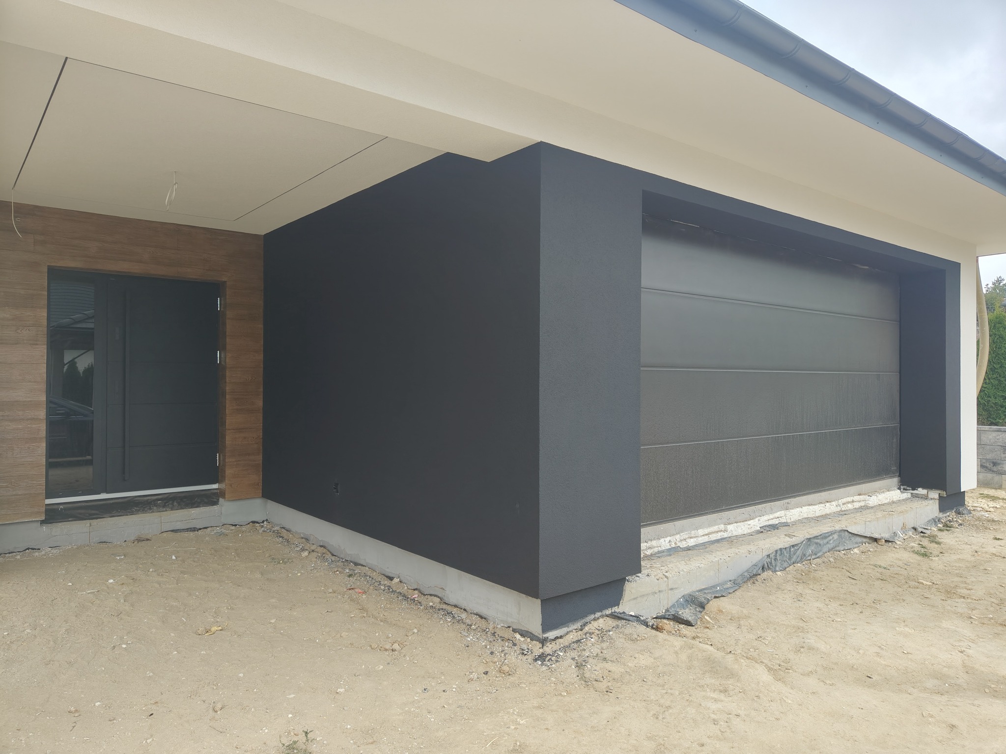 Nowoczesna elewacja domu z antracytową ścianą i bramą garażową. Widoczny fragment wejścia z drewnopodobną okładziną. Surowy teren wokół budynku.