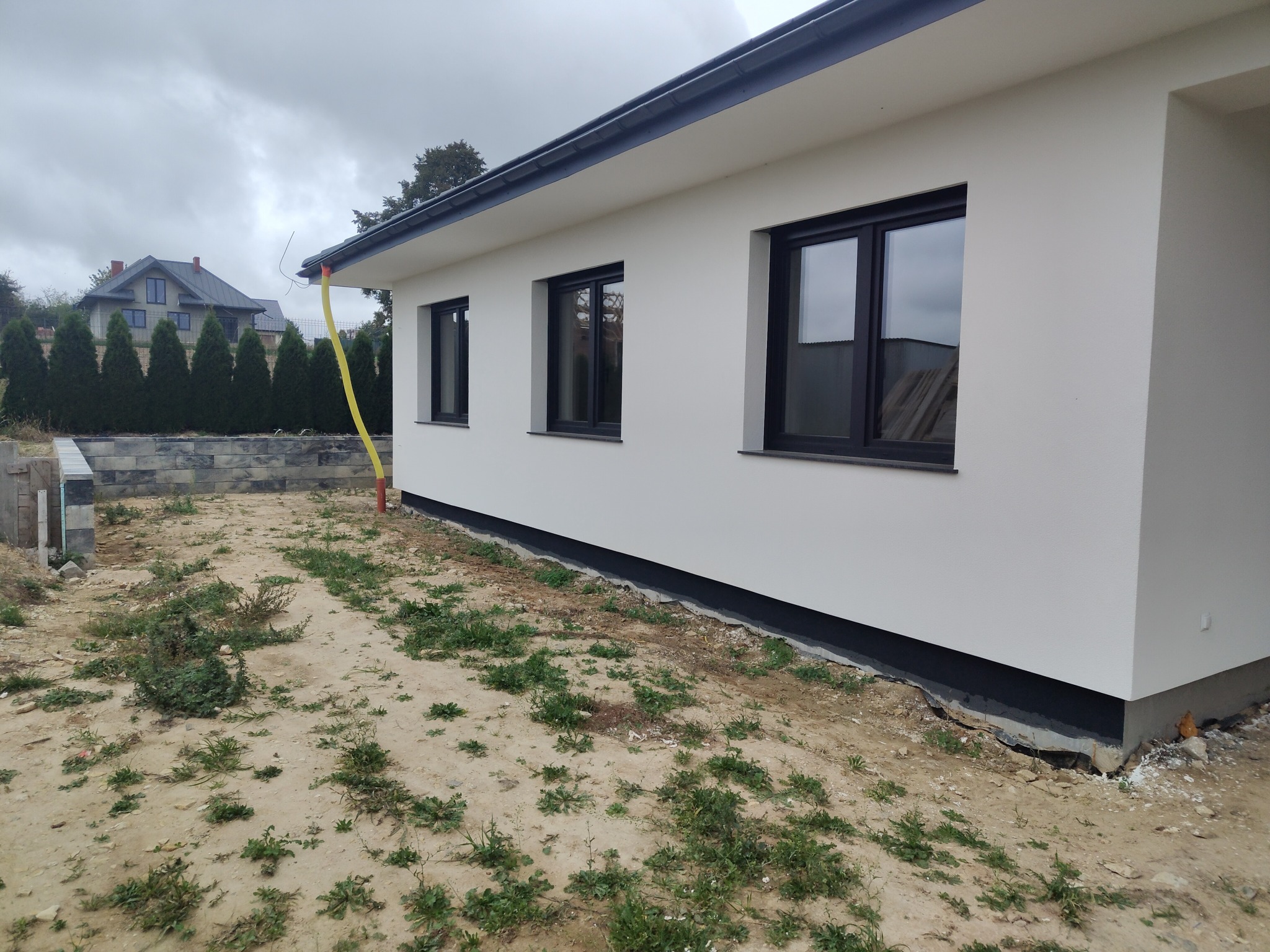 Biała elewacja domu jednorodzinnego z czarnymi oknami w Mełgwi. Widoczny fragment ogrodu i sąsiedni budynek. Ujęcie z boku, w pochmurny dzień.
