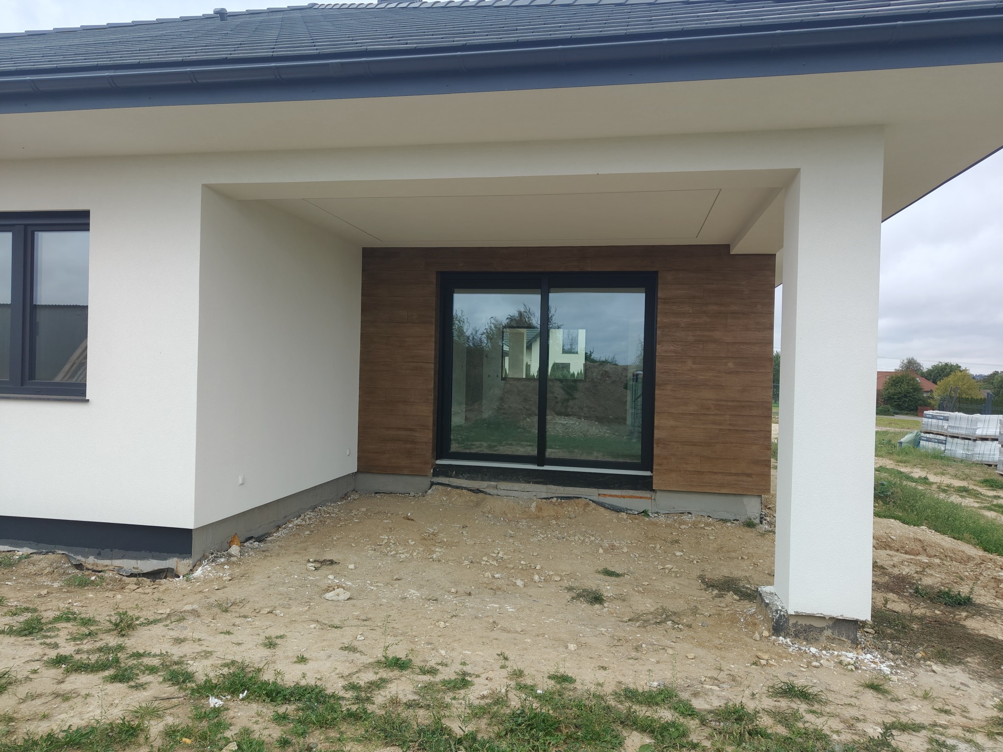 Nowoczesny dom z elewacją w odcieniach bieli i brązu. Widoczne duże okno przesuwne i fragment ogrodu. Elewacja z elementami dekoracyjnymi imitującymi drewno.