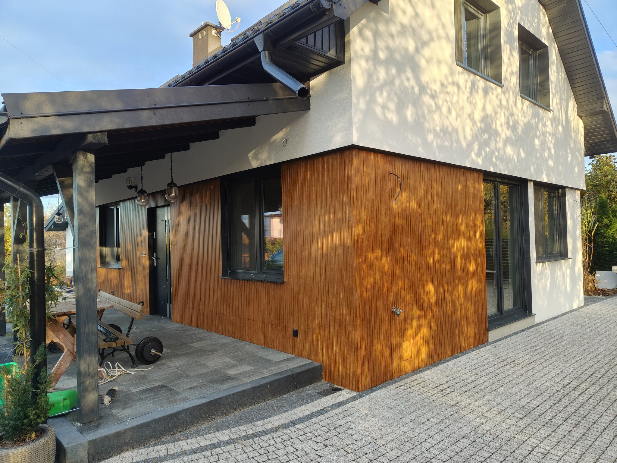 Nowoczesny dom z elewacją z drewnianych paneli i białym tynkiem, widok z zewnątrz, kostka brukowa na podjeździe, oświetlenie zewnętrzne, słoneczny dzień.