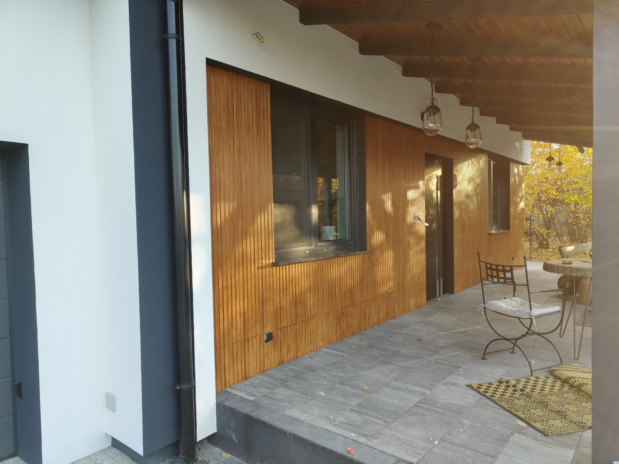 Nowoczesna elewacja domu z drewnianą okładziną i szarą stolarką okienną. Widoczny fragment tarasu z meblami ogrodowymi i jesienny krajobraz w tle.