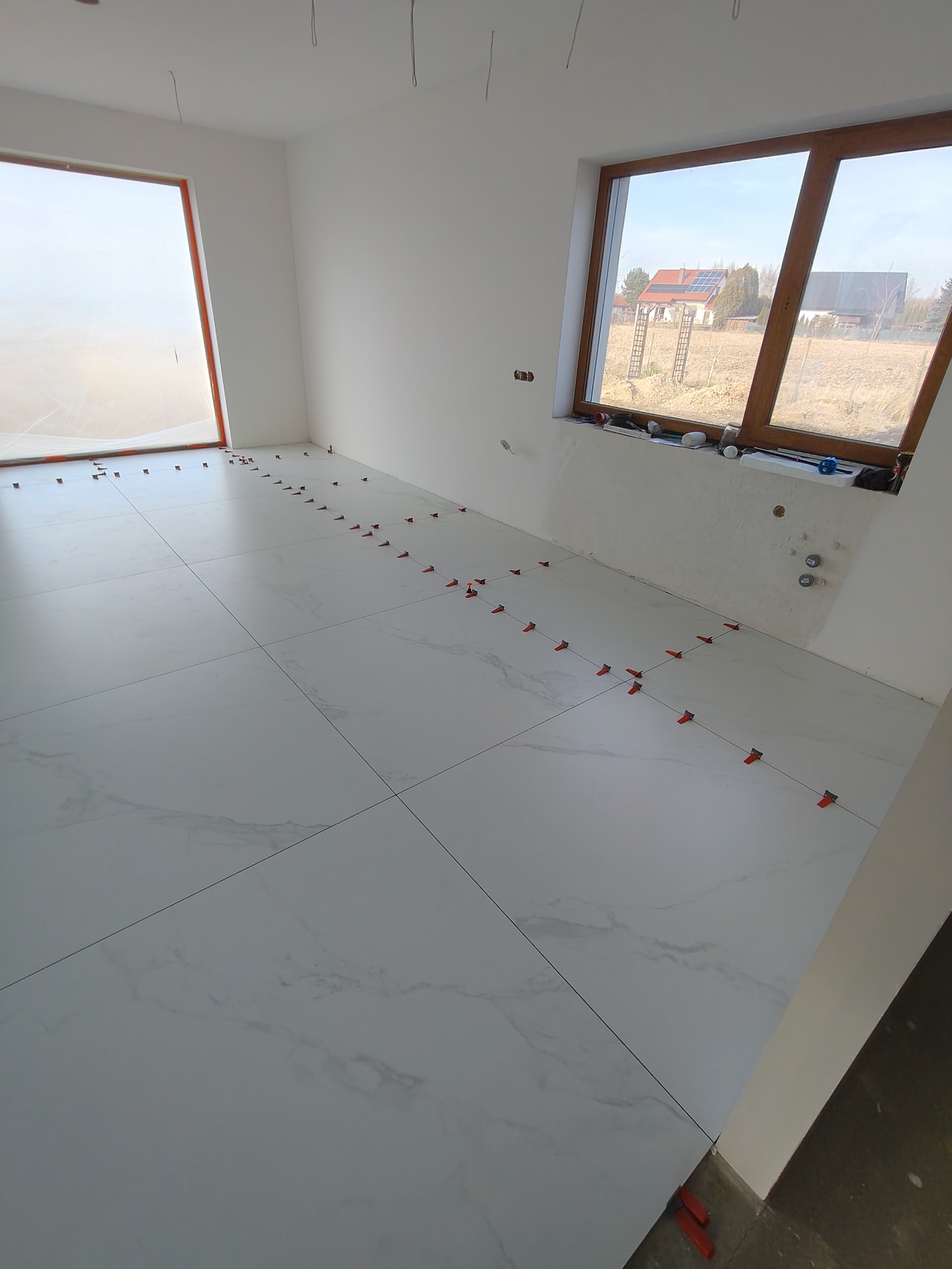 Układanie białych płytek podłogowych z marmurkowym wzorem w jasnym pomieszczeniu. Widoczne krzyżyki dystansowe i okno z widokiem na domy.