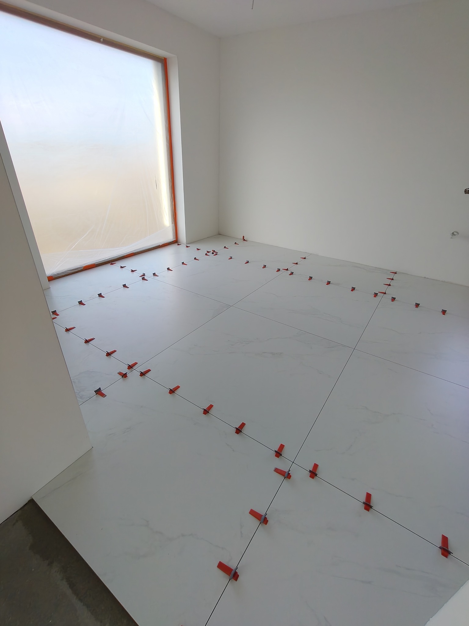 Białe płytki podłogowe w trakcie układania, z czerwonymi klipsami poziomującymi, w jasnym pomieszczeniu z oknem zabezpieczonym folią. Widok na ułożoną podłogę.