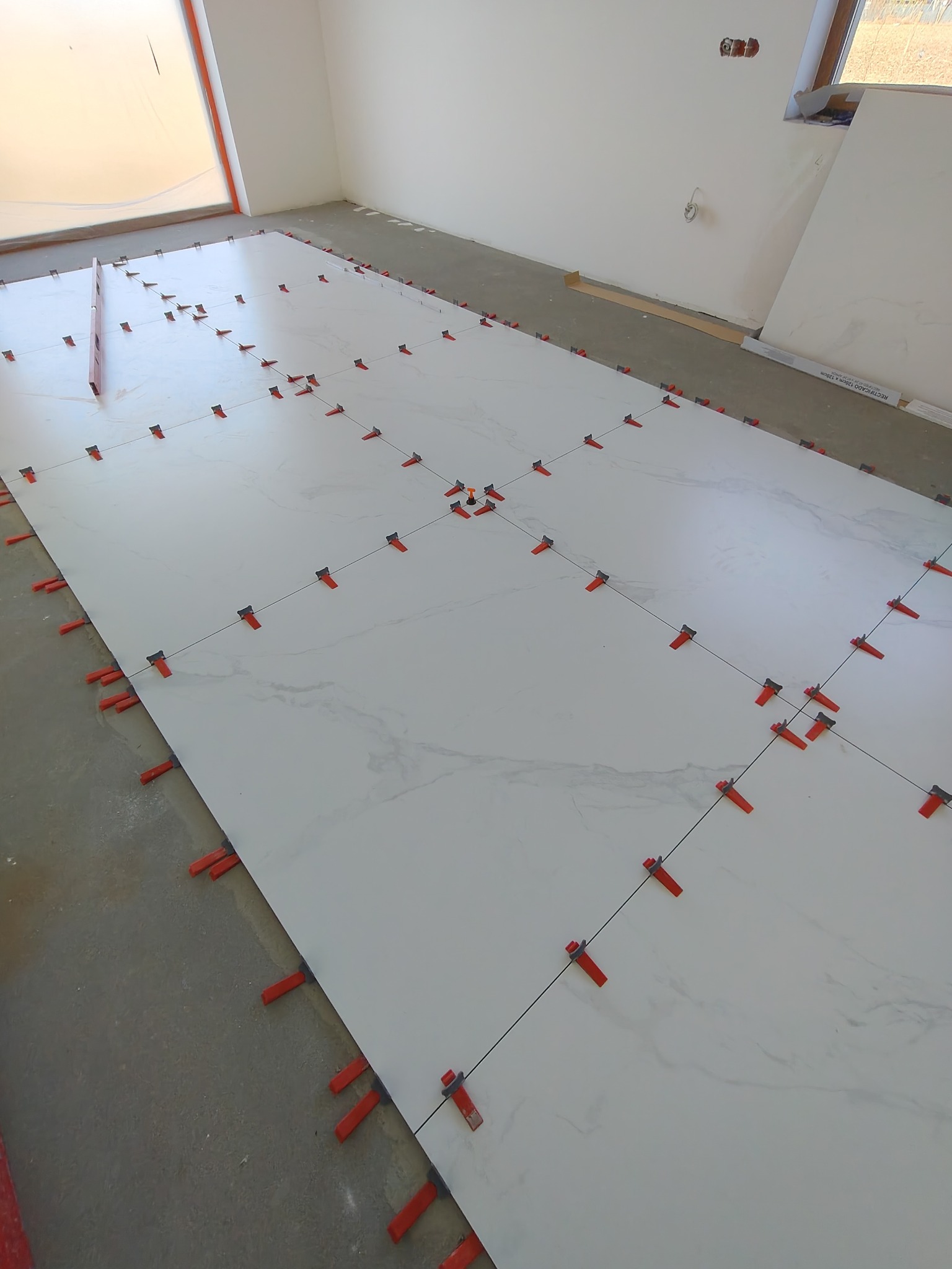 Układanie białych, marmurkowych płytek podłogowych z użyciem czerwonych klipsów poziomujących. Widoczny poziomica i fragment okna. Jasne wnętrze w trakcie prac wykończeniowych.