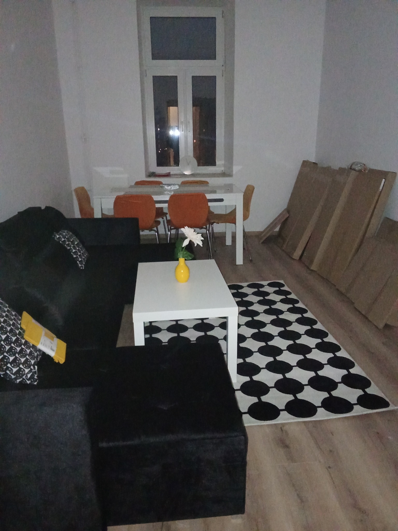 Umeblowane wnętrze pokoju z czarną sofą, białym stolikiem z wazonem, stołem z krzesłami i kartonami opartymi o ścianę. Widok na okno.