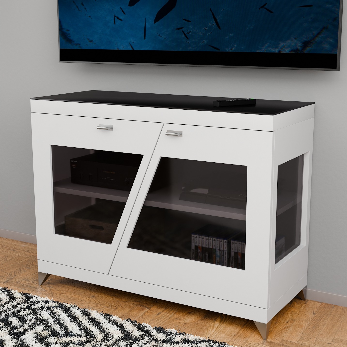 Biała szafka RTV z czarnym blatem i szklanymi drzwiczkami, na metalowych nóżkach. Na blacie leży pilot. W tle telewizor i dywan w geometryczne wzory.