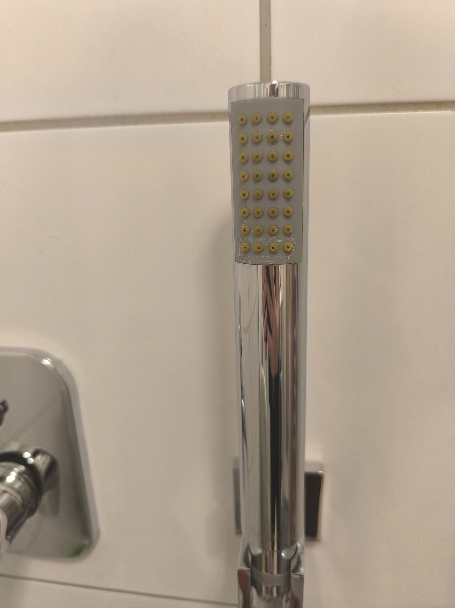 Błyszcząca, chromowana słuchawka prysznicowa z żółtymi dyszami, zamontowana na białej, kafelkowanej ścianie w łazience. Widoczny fragment baterii.