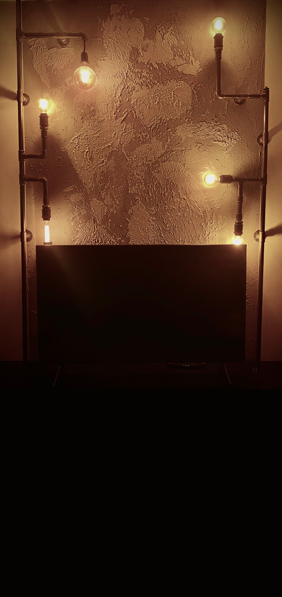 Industrialna lampa ścienna z rur i żarówek Edisona, zamontowana na tle ściany o fakturze betonu. Ciepłe światło tworzy unikalny klimat w pomieszczeniu.