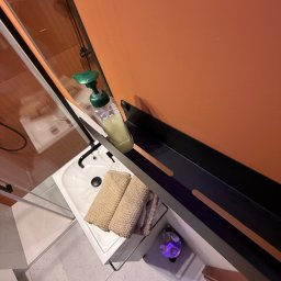 Aviendo sp. z o.o. - Umywalka w łazience z dozownikiem mydła, ręcznikami i czarną półką na tle pomarańczowej ściany. Widok z góry, oświetlenie punktowe.