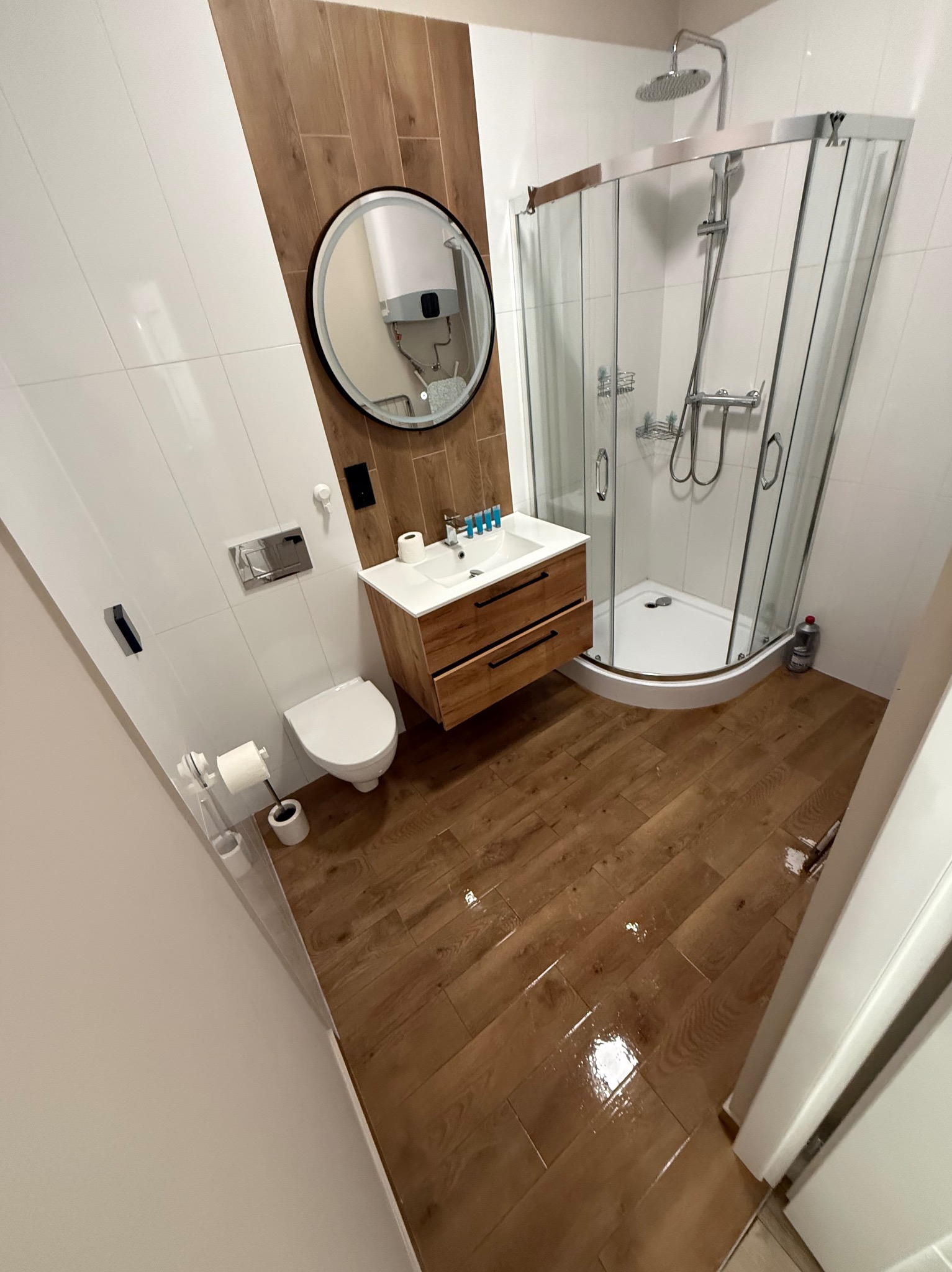 Nowoczesna łazienka z drewnianymi akcentami: podłoga, szafka pod umywalką i dekoracyjny panel. Kabina prysznicowa, toaleta i okrągłe lustro dopełniają wystrój.