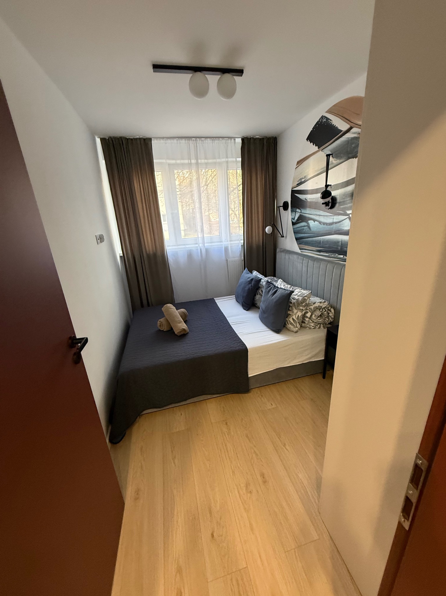 Przytulna sypialnia z łóżkiem, szarą pościelą i dekoracyjnym panelem ściennym. Widok na okno z brązowymi zasłonami i drewnianą podłogę.