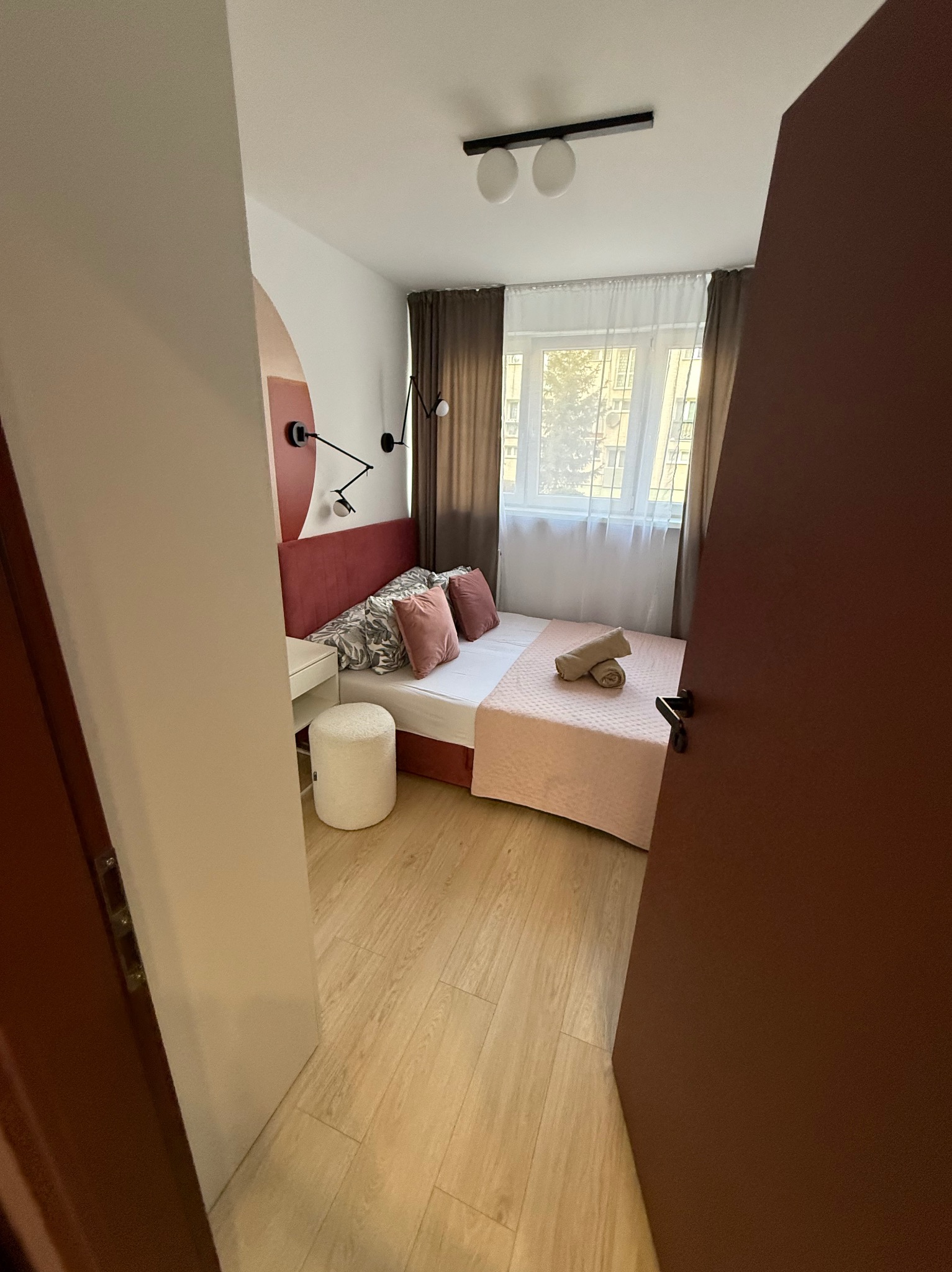 Ustawne wnętrze sypialni z łóżkiem, stolikiem i pufą, widziane zza uchylonych, brązowych drzwi. Stonowana kolorystyka, nowoczesne lampy ścienne.