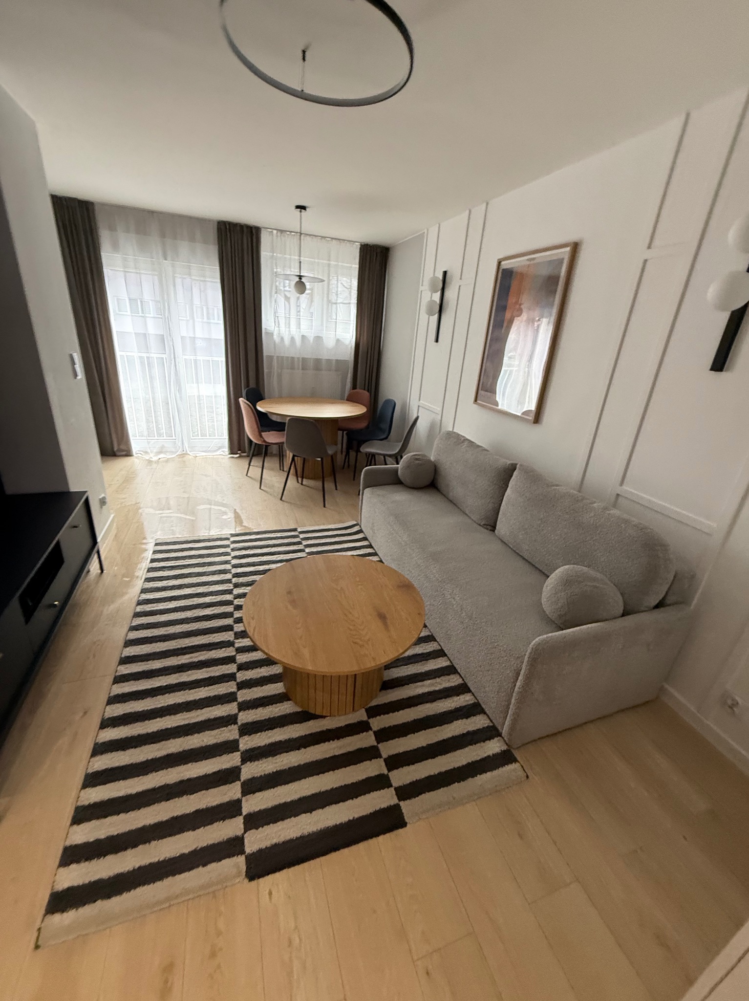 Jasne wnętrze salonu z szarą sofą, okrągłym stolikiem kawowym i dywanem w czarno-białe pasy. W tle stół z krzesłami i okno z zasłonami.